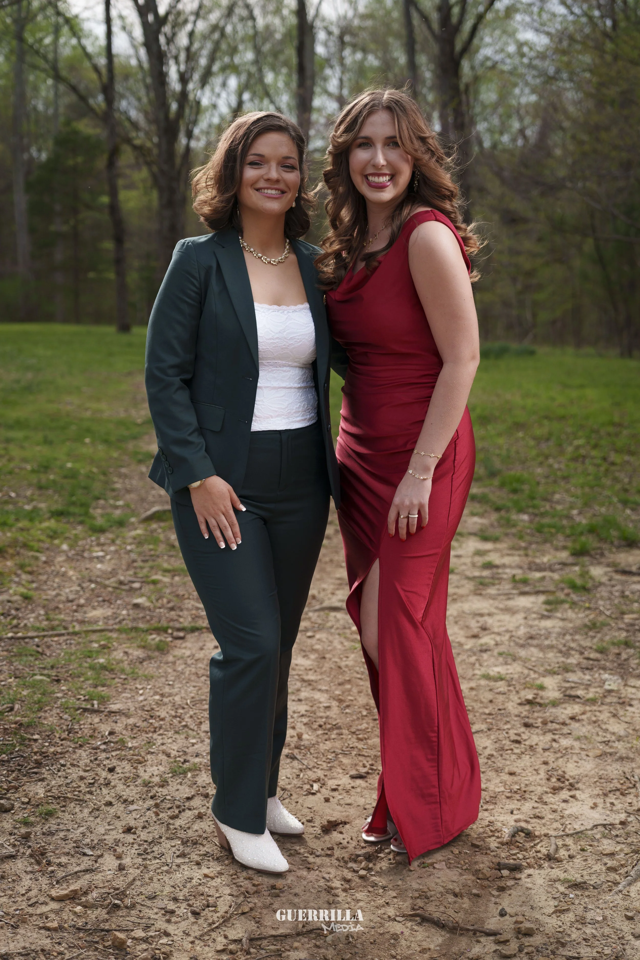 Two women standing outdoors on a dirt path, smiling at the camera, with trees and greenery in the background.