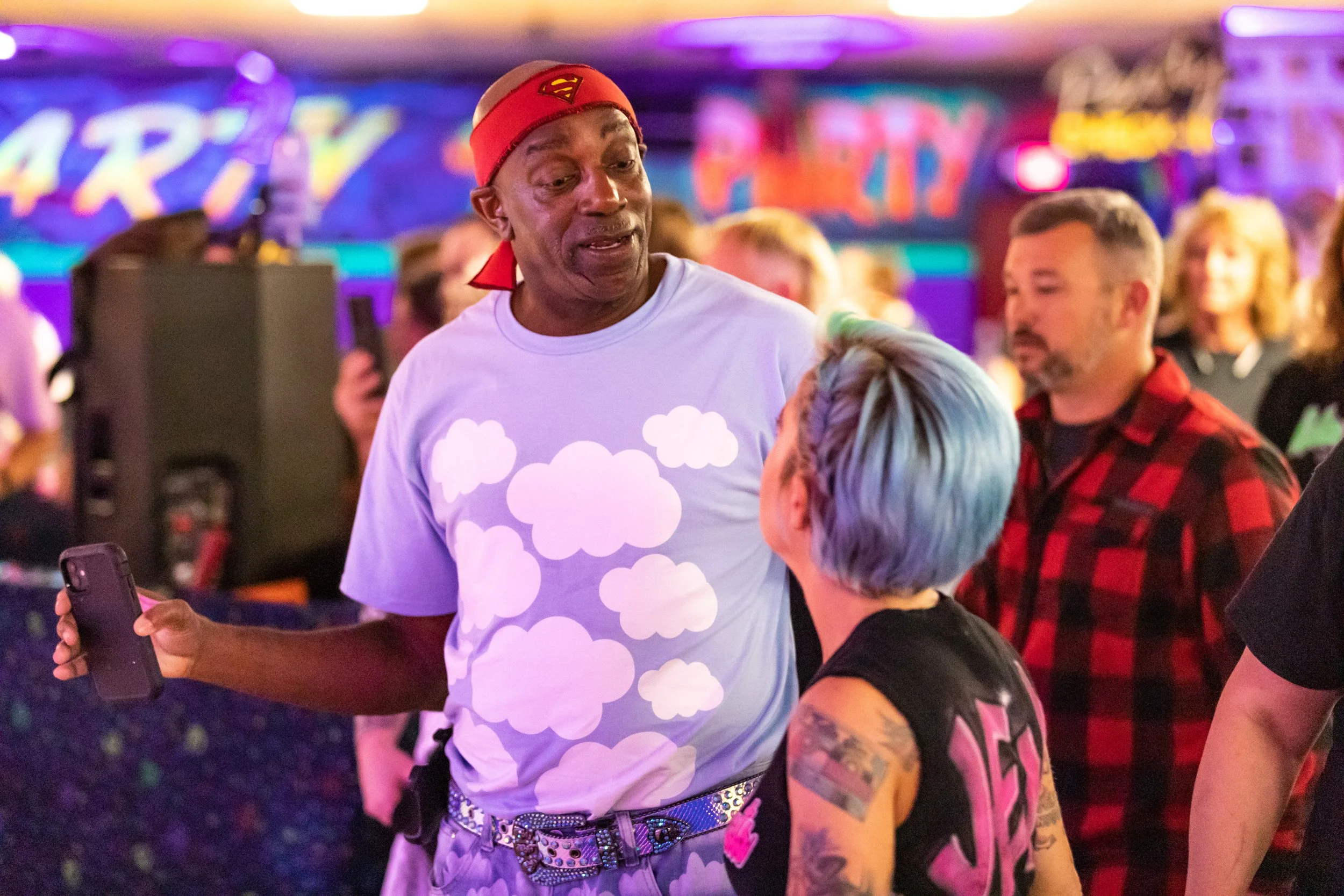 A man wearing a red band and a purple shirt with cloud patterns talking to a woman with colorful hair in a crowded party setting with neon signs and colorful lighting.
