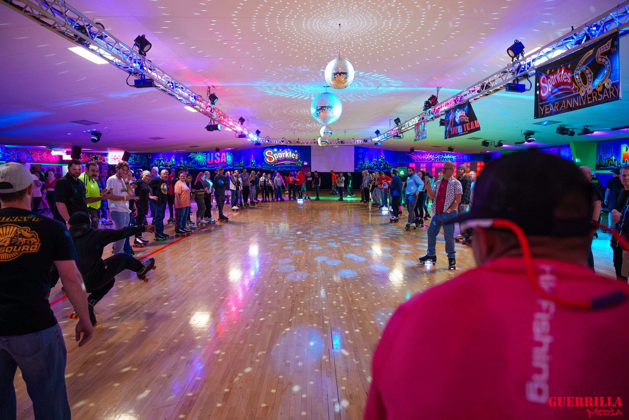 A roller skating rink filled with people skating and socializing, decorated with colorful neon signs and lighting for a celebration, with disco balls hanging from the ceiling and a wooden floor.