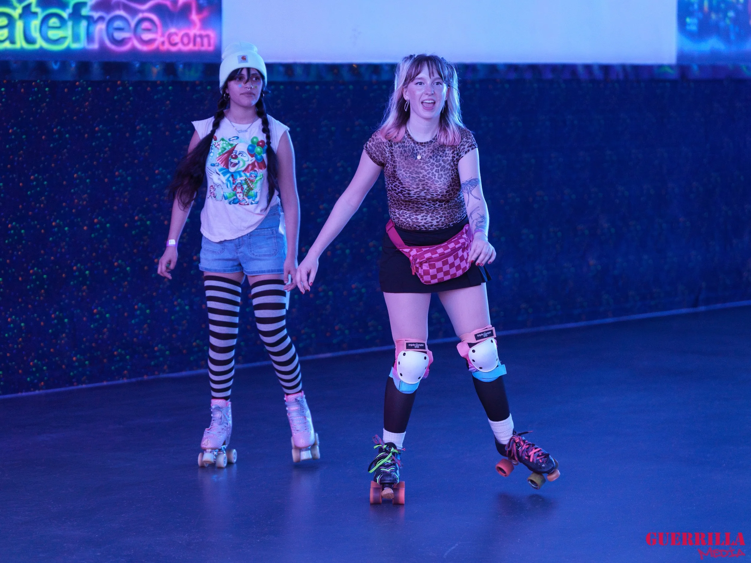 Two young women roller skating in an indoor rink, one with a pink fanny pack and knee pads, the other wearing striped tights and a beanie, with a colorful sign in the background.
