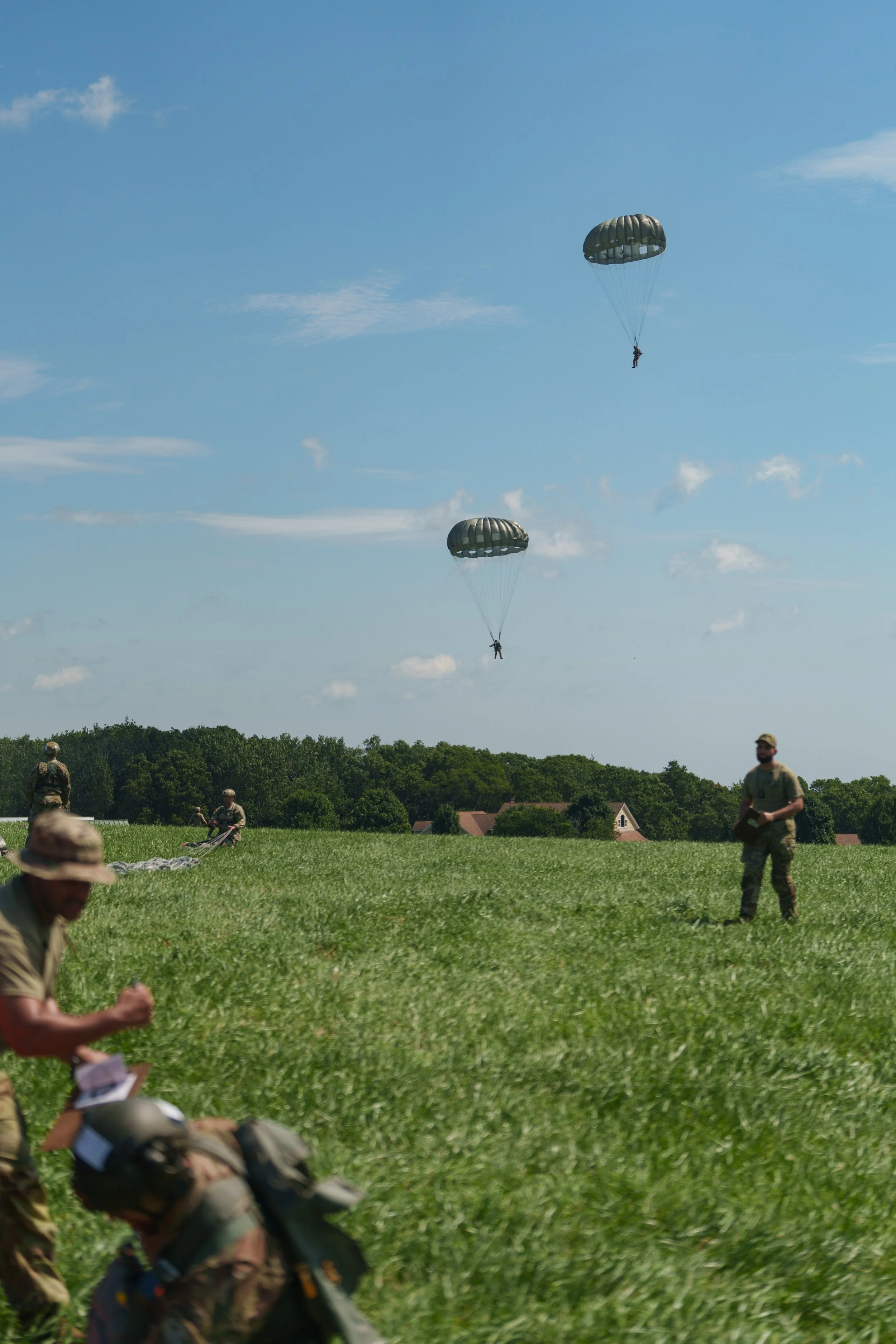 Two parachuters descending from the sky while military personnel and equipment are on the ground in a grassy field.