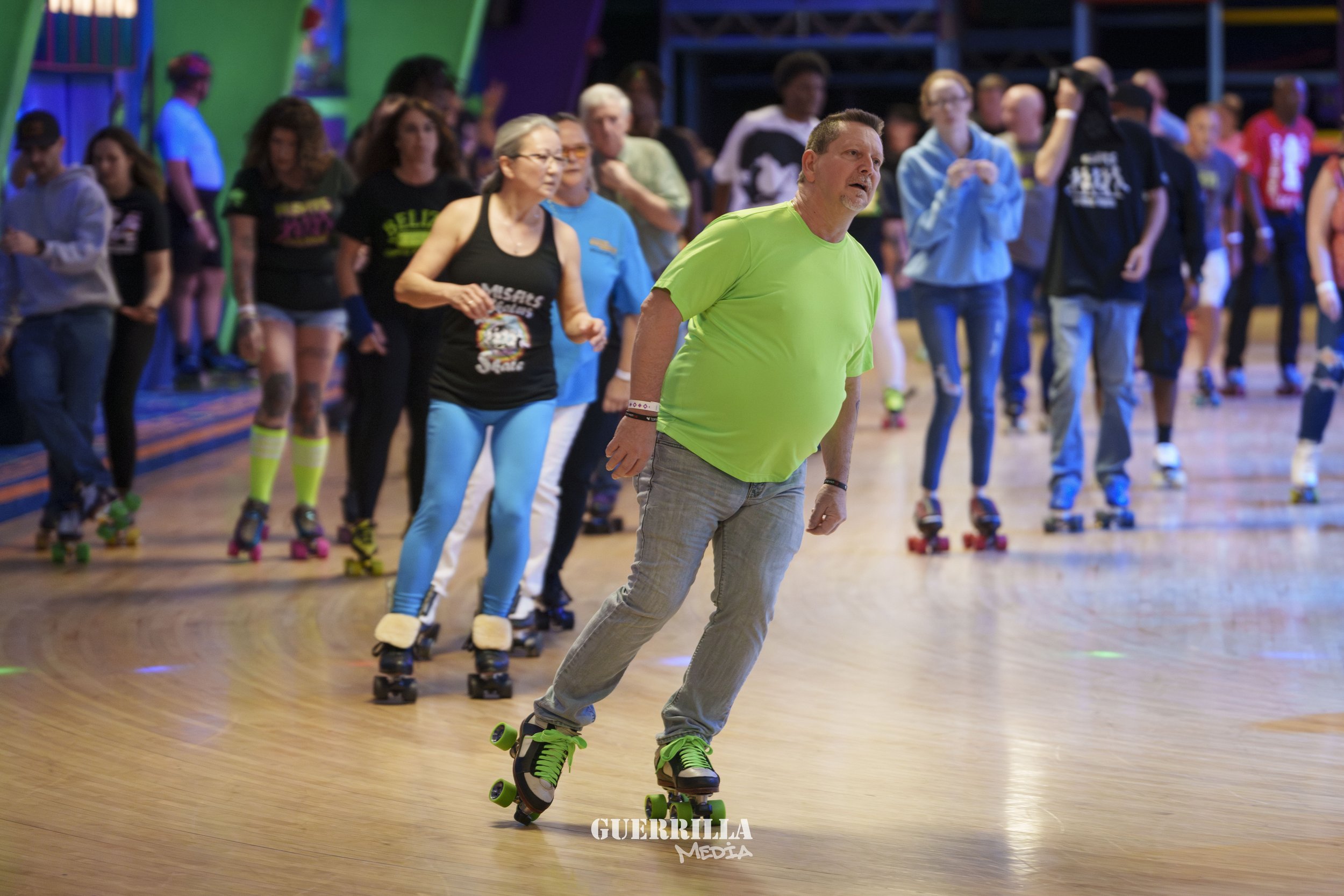 People roller skating indoors, with a man in a bright green shirt in the foreground and others in the background, some of whom are wearing roller skates and athletic clothing.