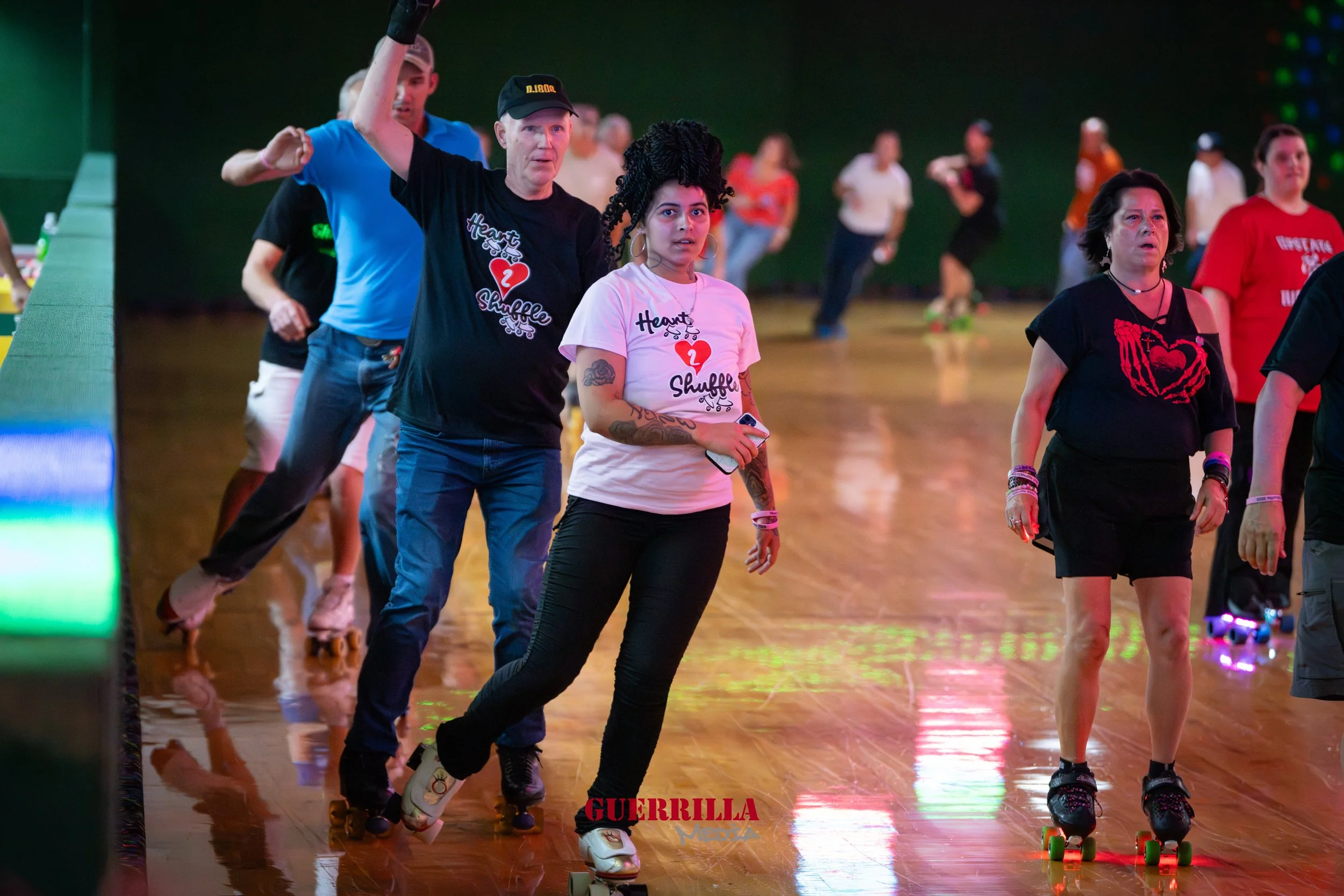 People roller skating indoors, wearing casual clothes, with a woman in the foreground holding a phone and wearing a white t-shirt with text. The background is blurred with more skaters and colorful lights.