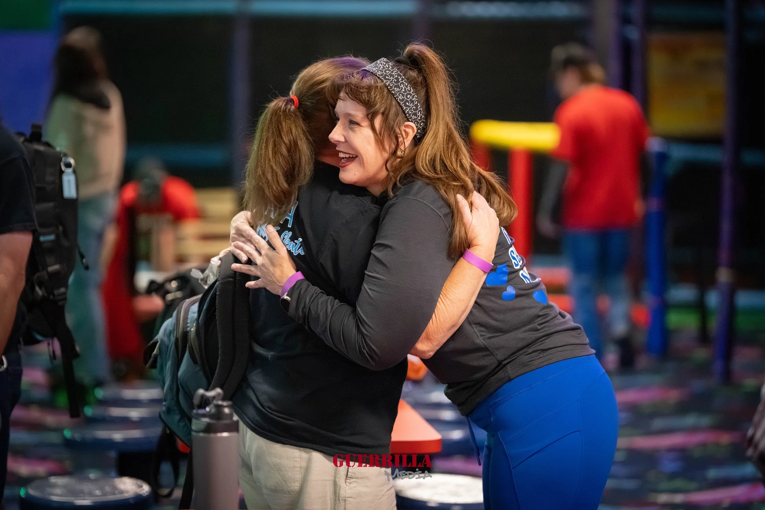 Two women hugging and smiling in a brightly lit indoor setting, with colorful decor and other people in the background.