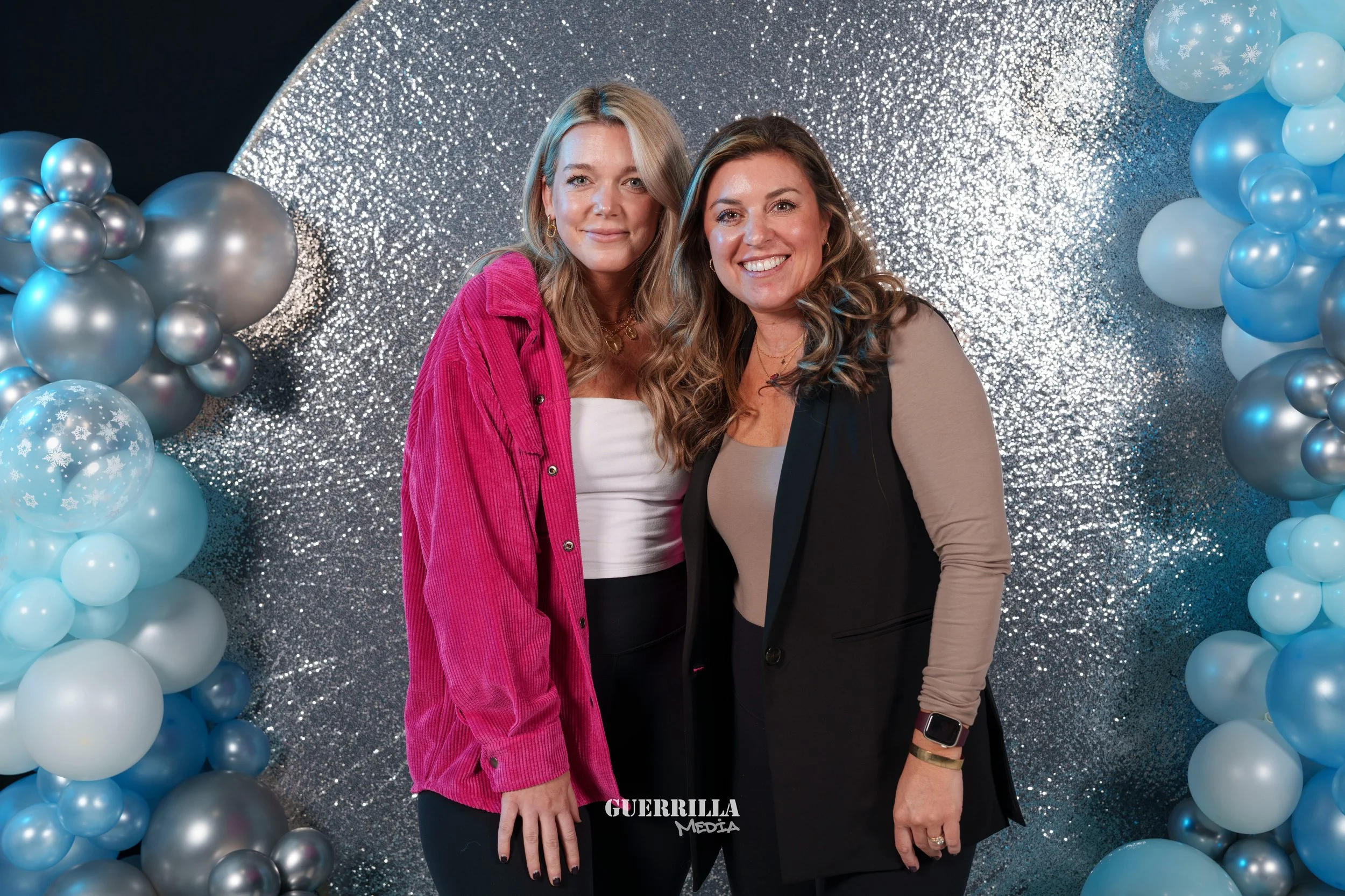 Two women smiling and posing together in front of a glittery silver and blue balloon backdrop at an event.