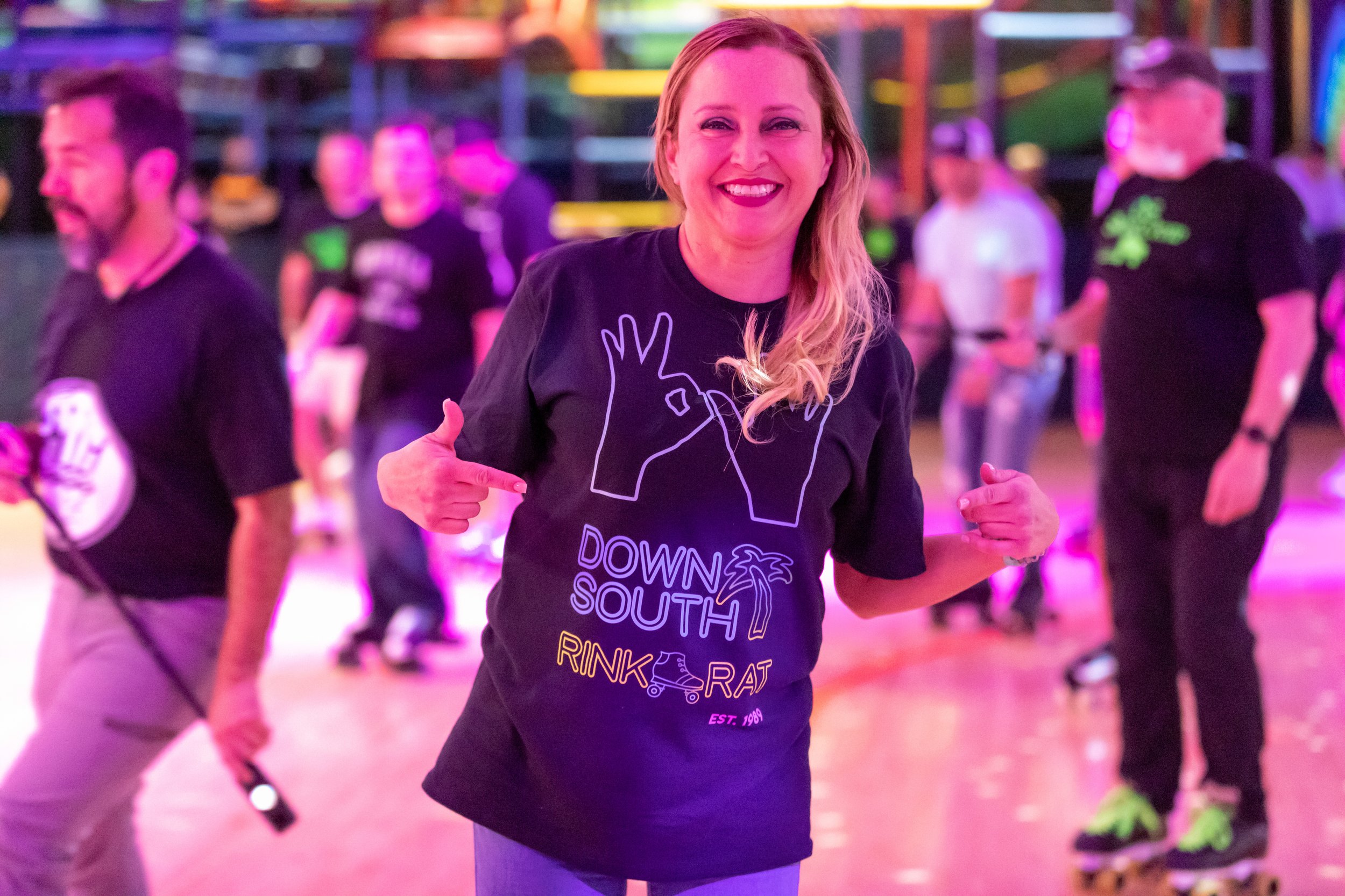 Woman smiling and pointing at her black T-shirt with a graphic of two hands and the words 'Down South Rink Rat' at a roller skating rink with other people in the background.