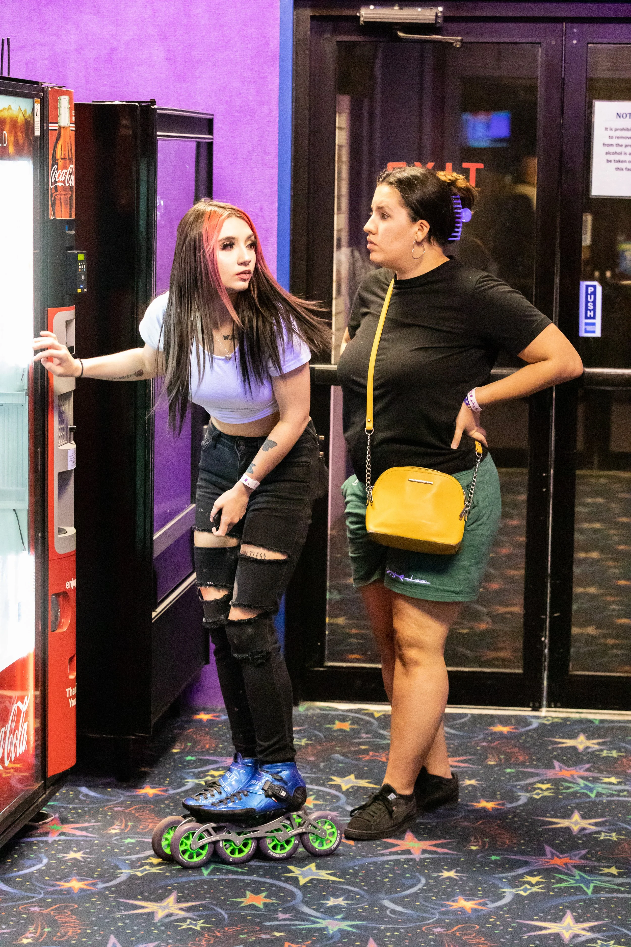 Two women at an arcade, with one on roller skates and talking to the other, near a vending machine, in a colorful, star-patterned floor.