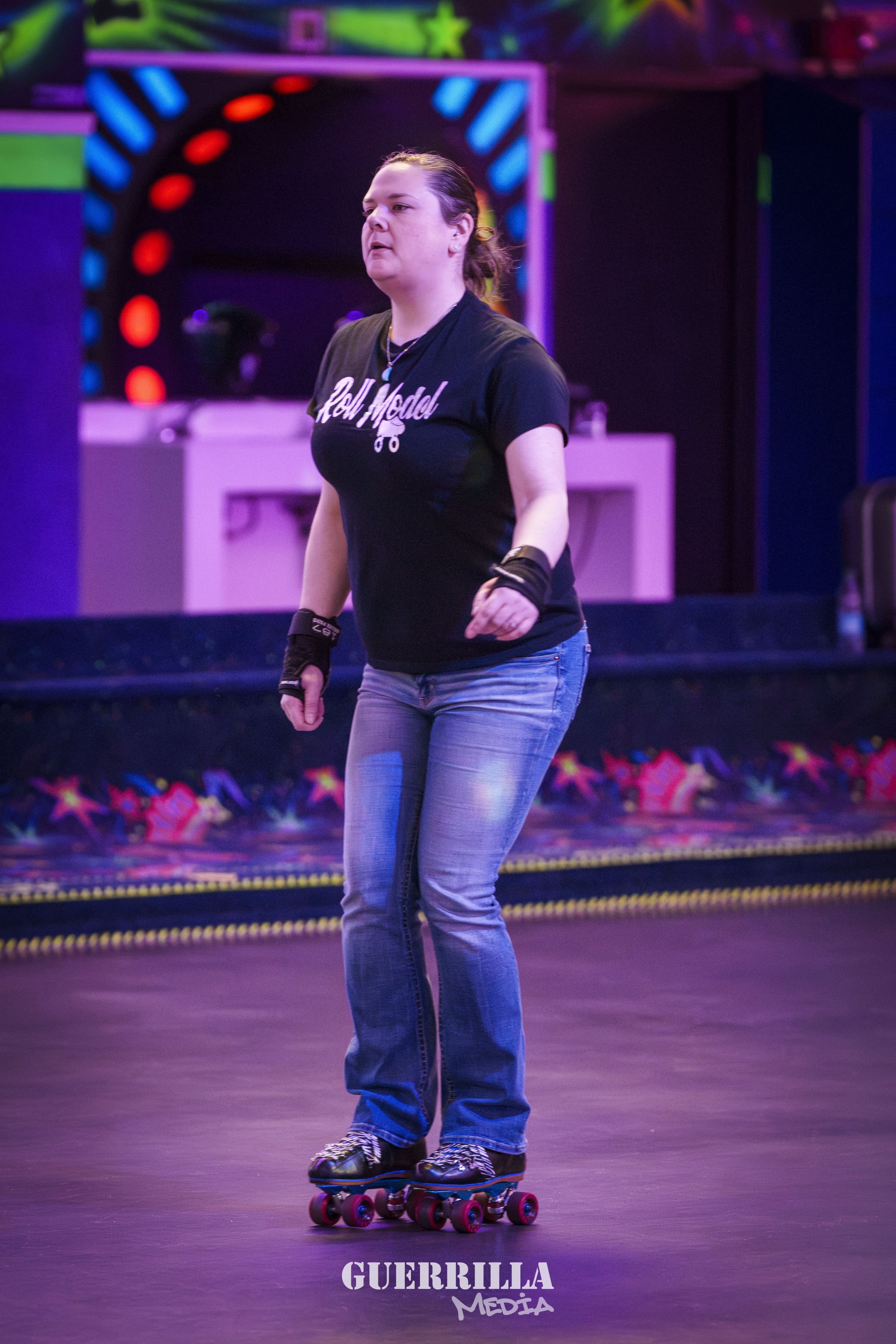 A woman roller skating inside a venue with purple and colorful lights in the background, wearing a black t-shirt, jeans, and protective gear on her wrists.