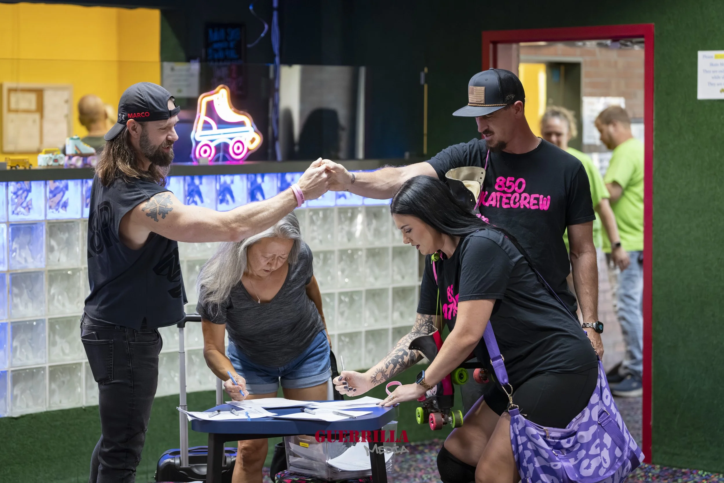 A group of people at an indoor skating rink, with one man giving a handshake to another man, while a woman is signing papers on a table. The woman has tattoos and is wearing roller skates, and there are neon lights in the background.