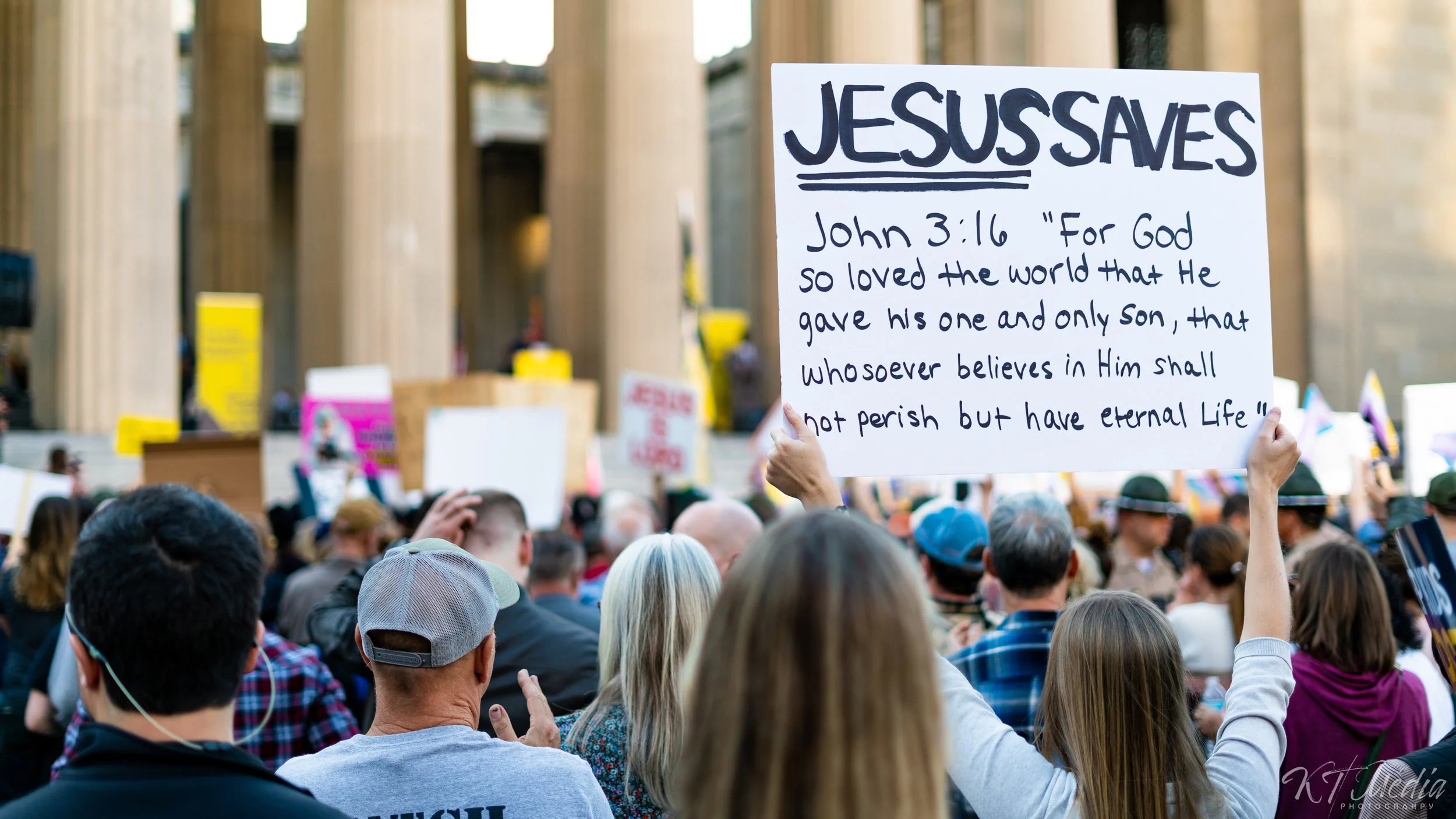 A crowd of people at a protest or rally, with one person holding a large sign referencing John 3:16 from the Bible.
