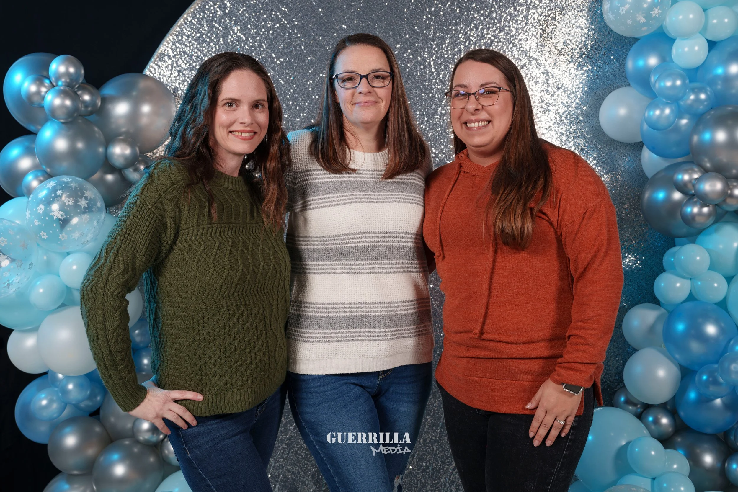 Three women standing together in front of a glittery silver and blue balloon background, smiling at the camera.