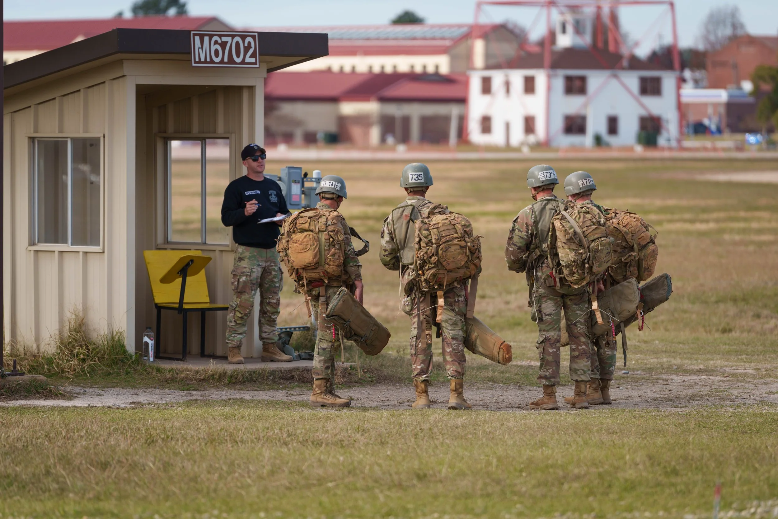 Military soldiers in camouflage uniforms and helmets standing in line with backpacks, receiving instructions from a supervisor during a training exercise at a military base.