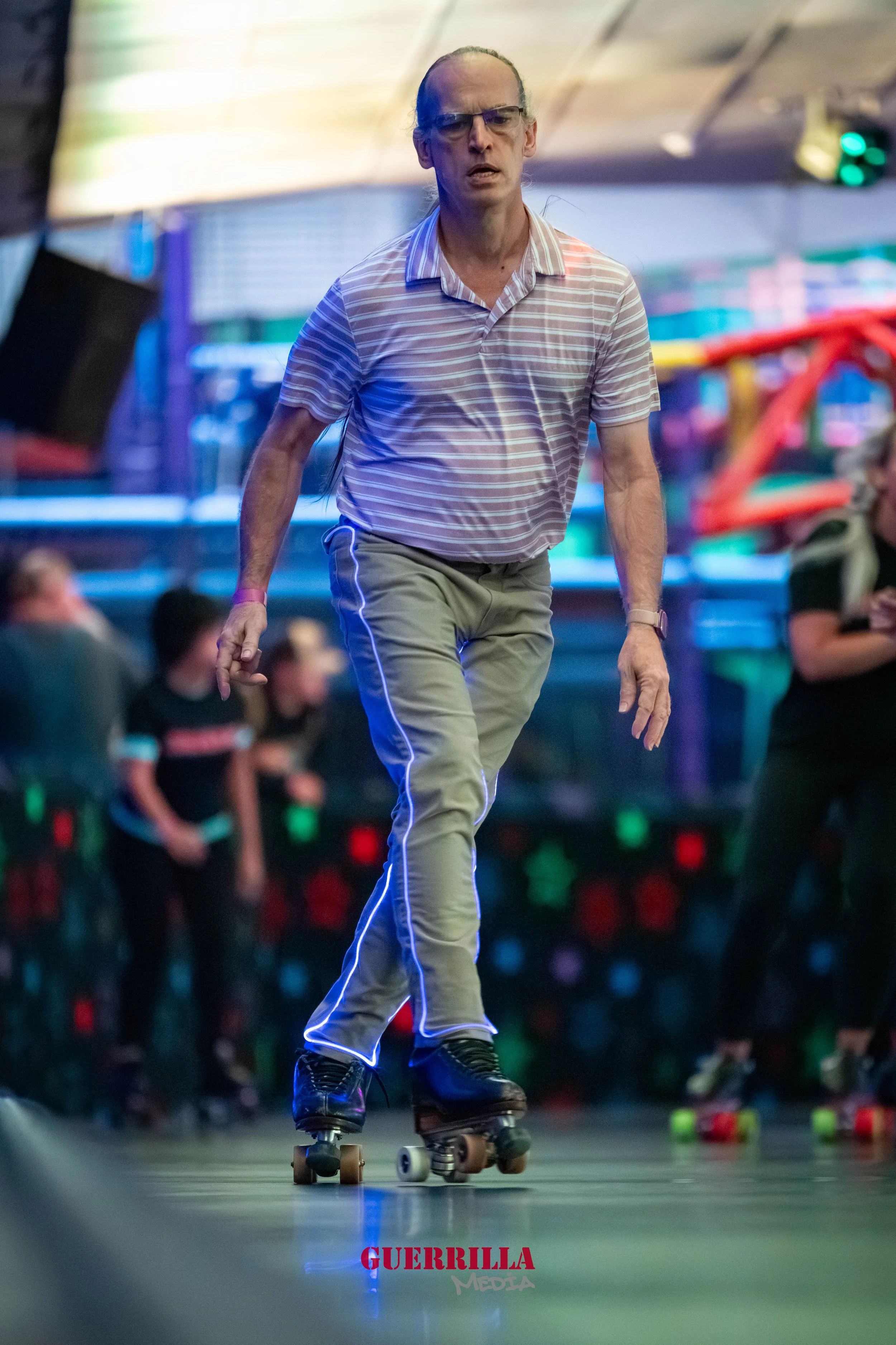 A man with glasses, wearing a striped polo shirt and beige pants, rollerblading indoors at an entertainment venue.