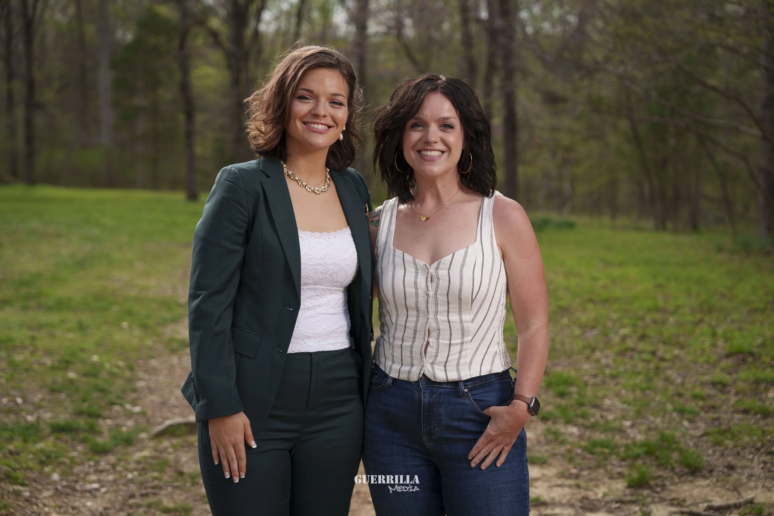 Two women smiling and standing close together outdoors in a wooded area with green grass and trees in the background.