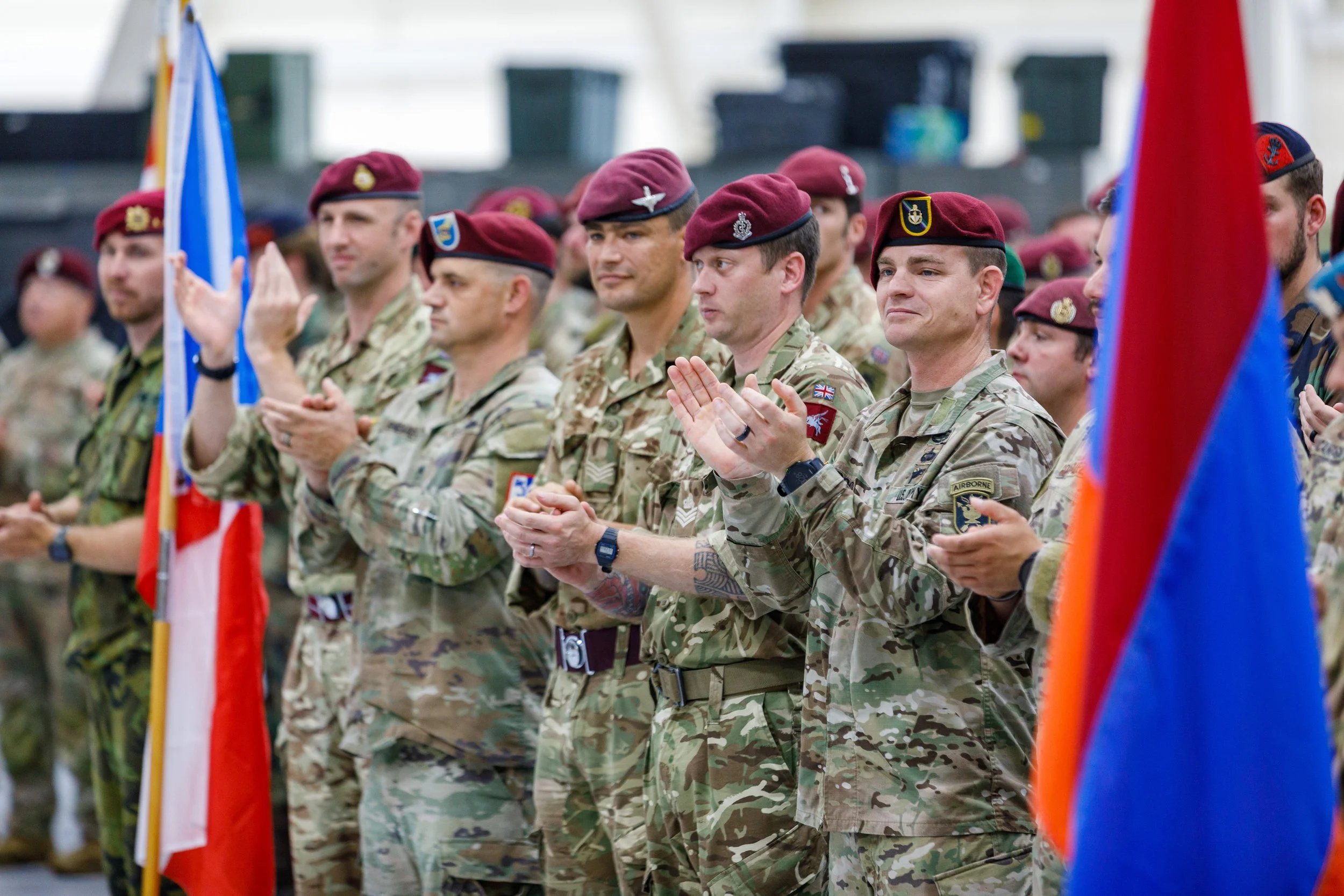 Military personnel in camouflage uniforms and maroon berets, standing and clapping during a ceremony, some holding flags, in an outdoor setting.