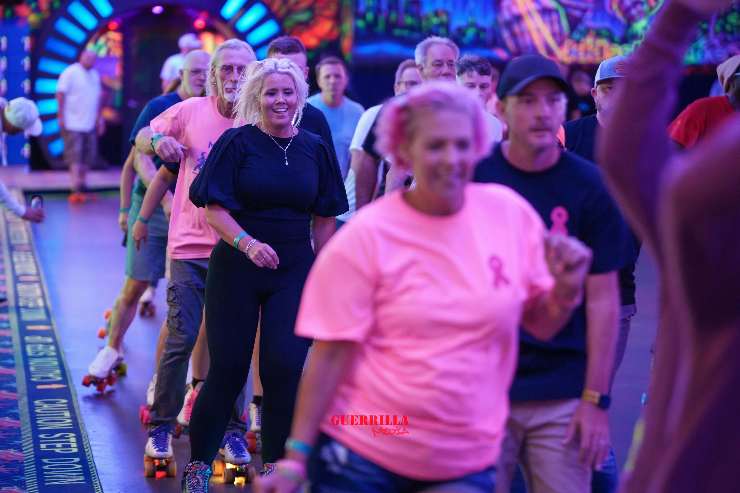 A group of people roller skating in an indoor rink with colorful neon lights in the background. Some are smiling and wearing pink ribbons. The group appears to be participating in a roller skating event or rally.