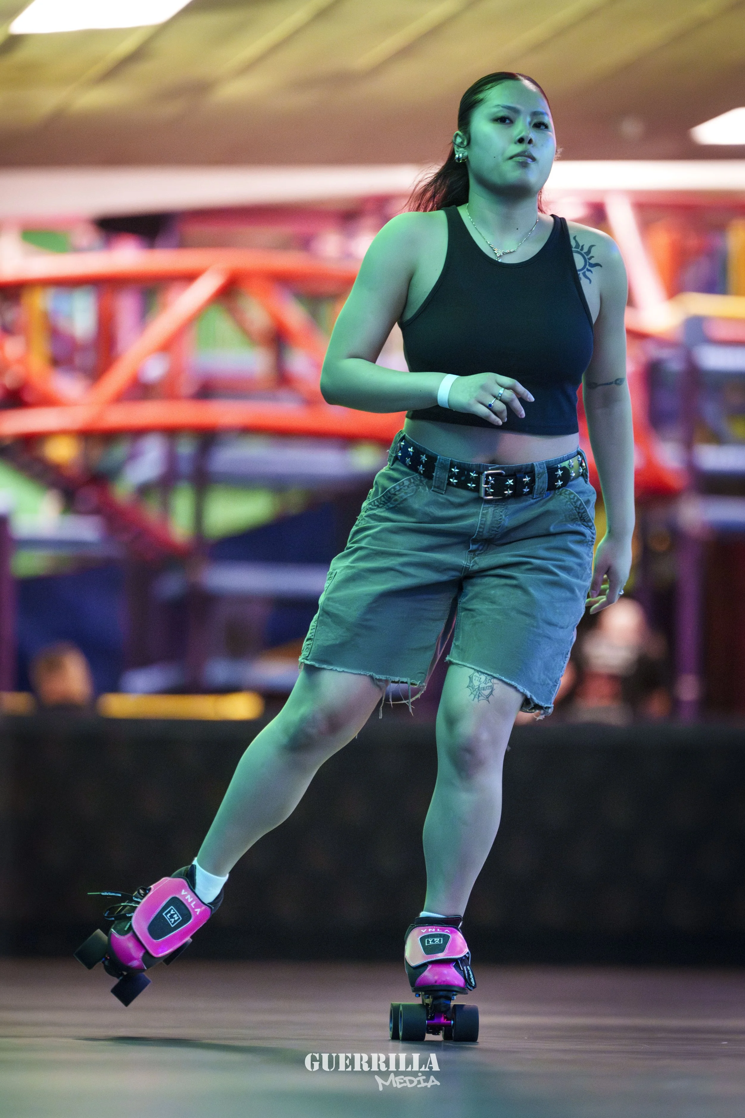 A woman in a black crop top and ripped shorts roller skating in an indoor roller rink, with colorful lights and blurred background.