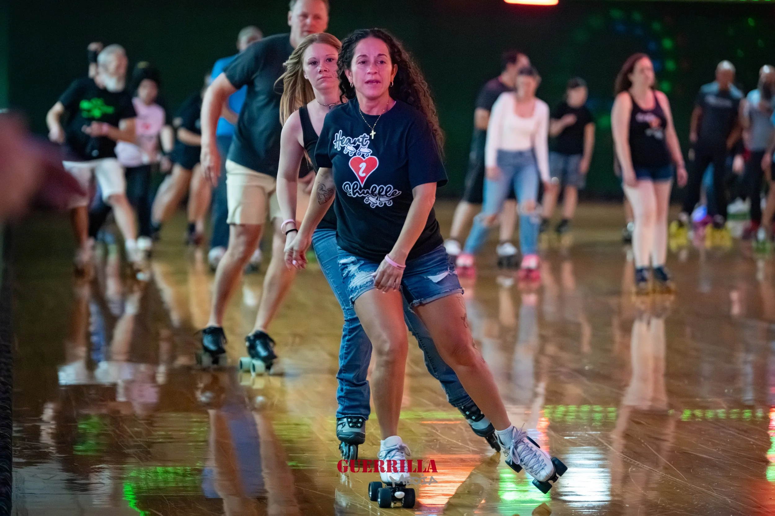 Multiple people roller skating indoors on a polished wooden floor, with a woman in the foreground wearing a black T-shirt and denim shorts, and others in the background.