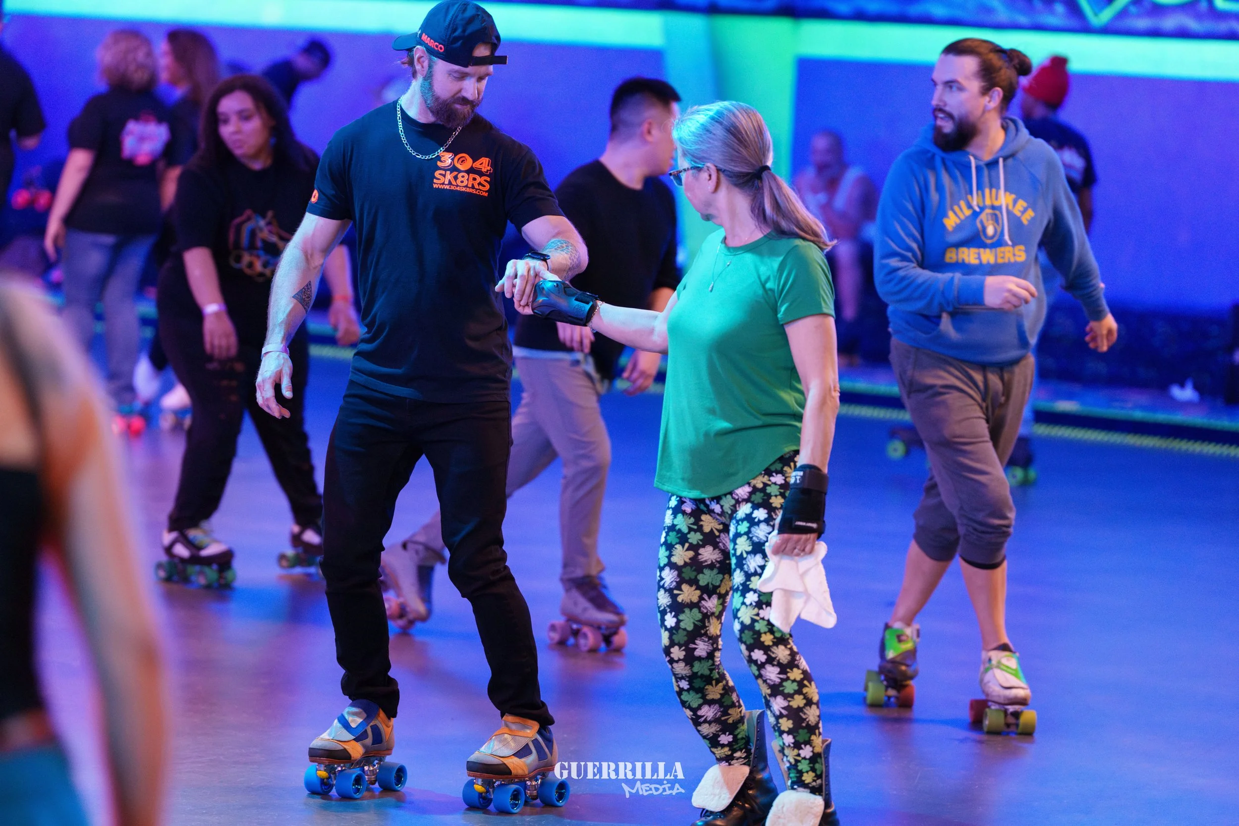 People roller skating in an indoor rink with colorful lighting. A man in a black T-shirt and a woman in a green shirt are holding hands, with the woman holding a cloth in her other hand. Other skaters are visible in the background.