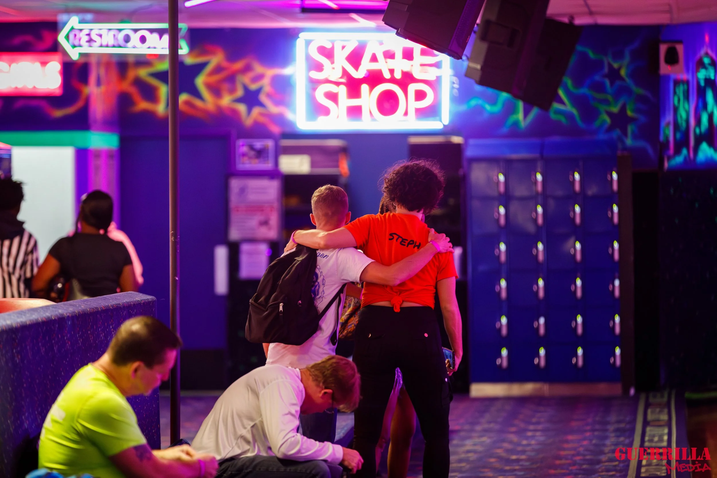 People sitting and standing in a neon-lit roller skating rink. Two people are hugging near the skate shop entrance, while others are seated or standing nearby. Bright neon signs and locker lockers are visible in the background.