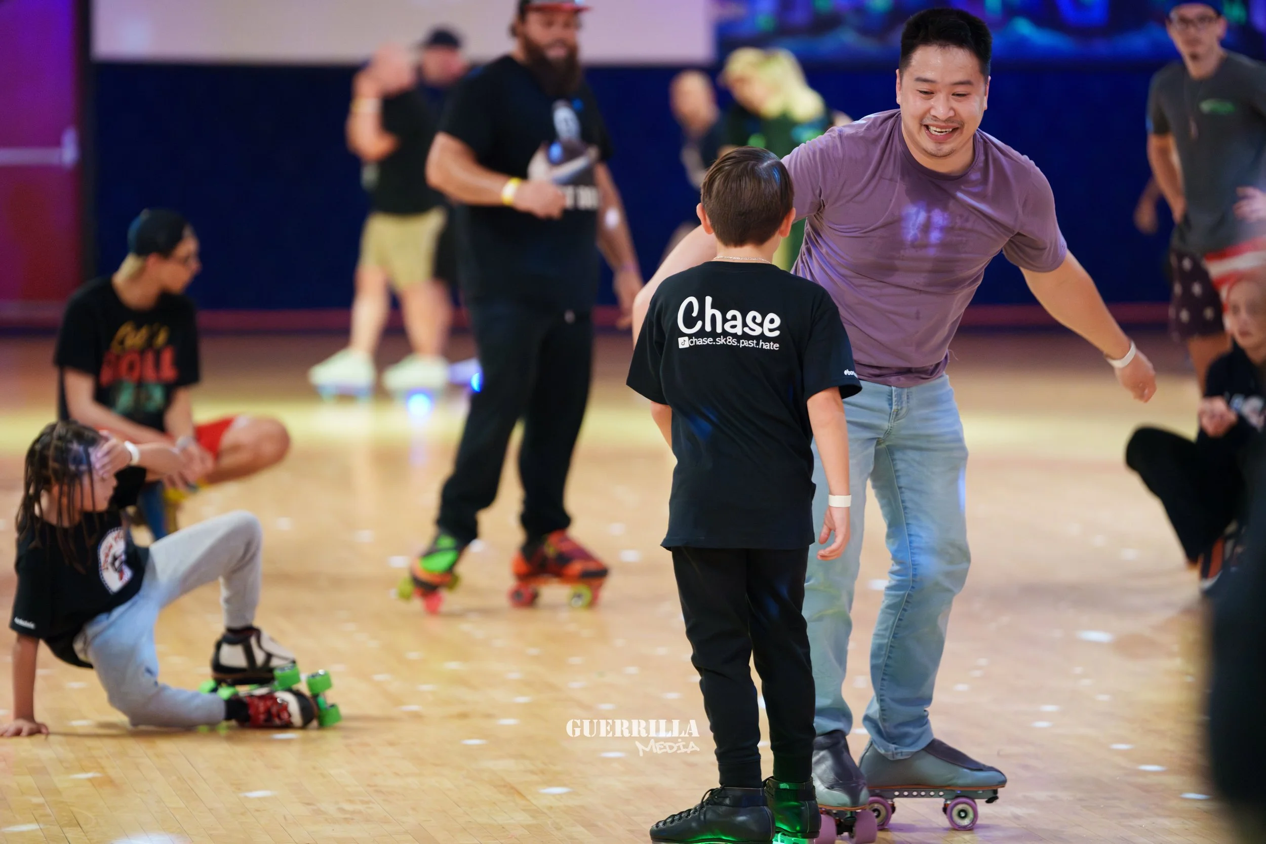A group of people roller skating indoors, with some sitting and others standing and skating, and a man smiling and talking to a young boy who is wearing a black shirt with 'Chase' printed on the back.