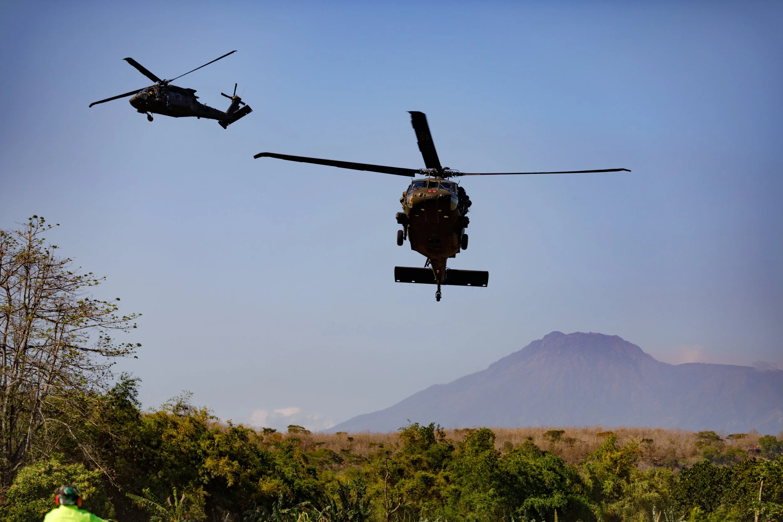 Two military helicopters flying over a landscape with trees and a mountain in the background.