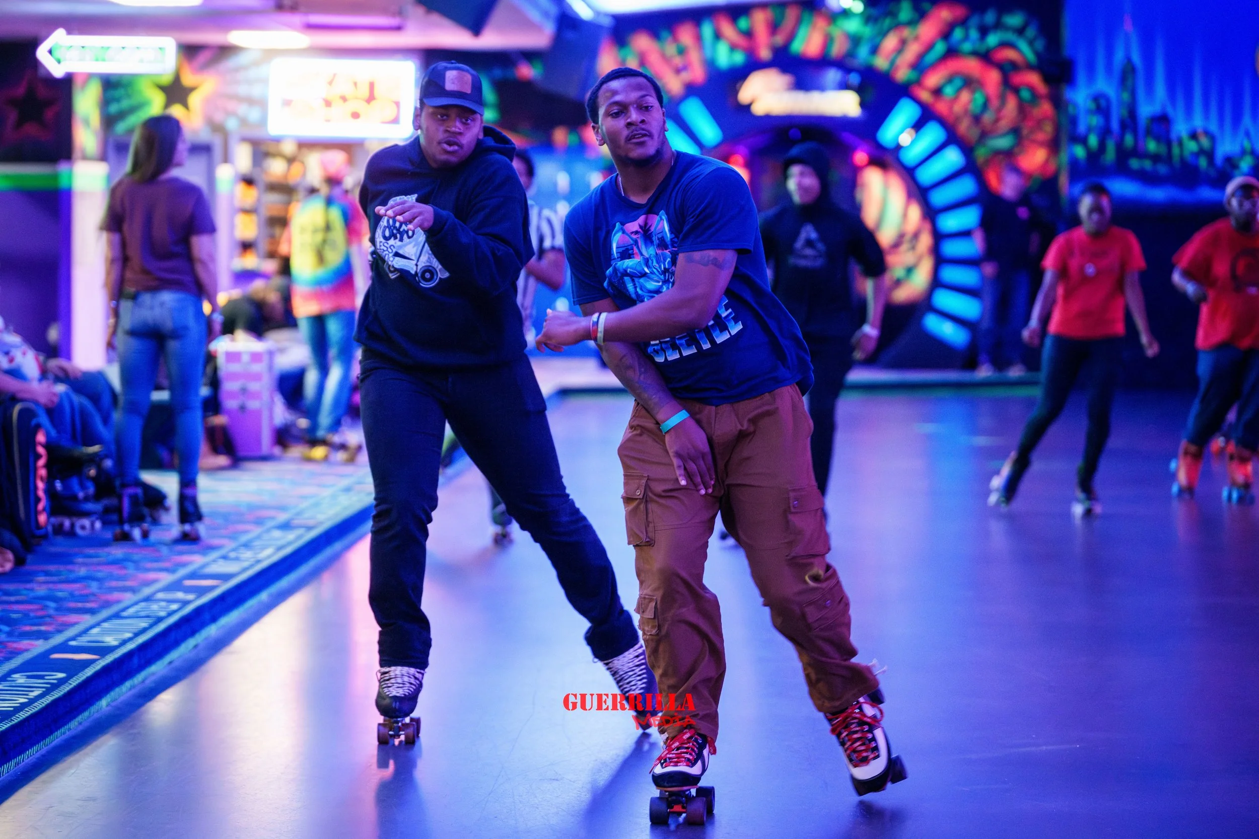 Two young men roller skating in an indoor skating rink with neon lights and colorful wall art, with other skaters and people in the background.