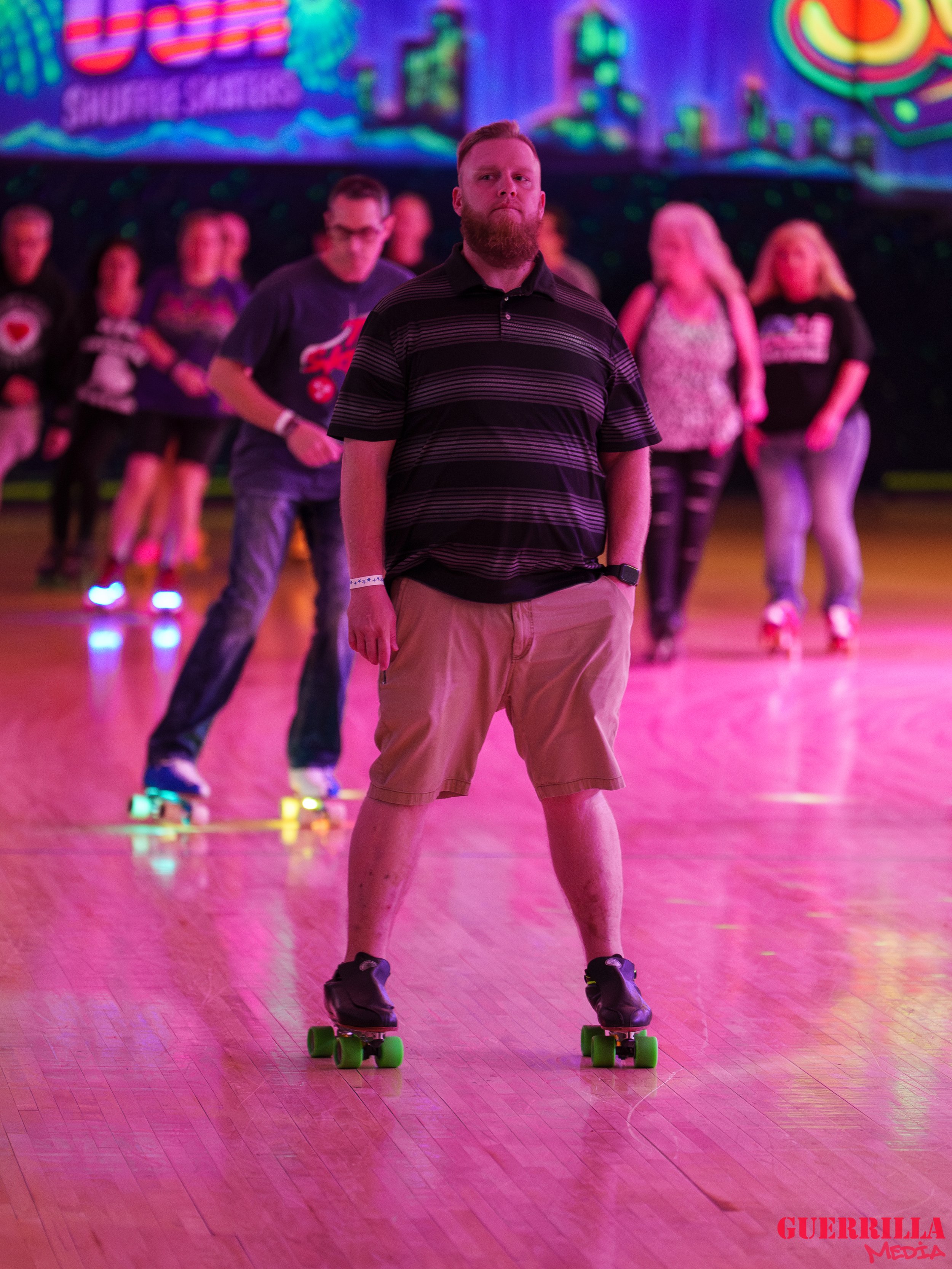 Man with a beard wearing a black striped polo and khaki shorts on roller skates at an indoor skating rink with pink lighting, with a group of people skating in the background.