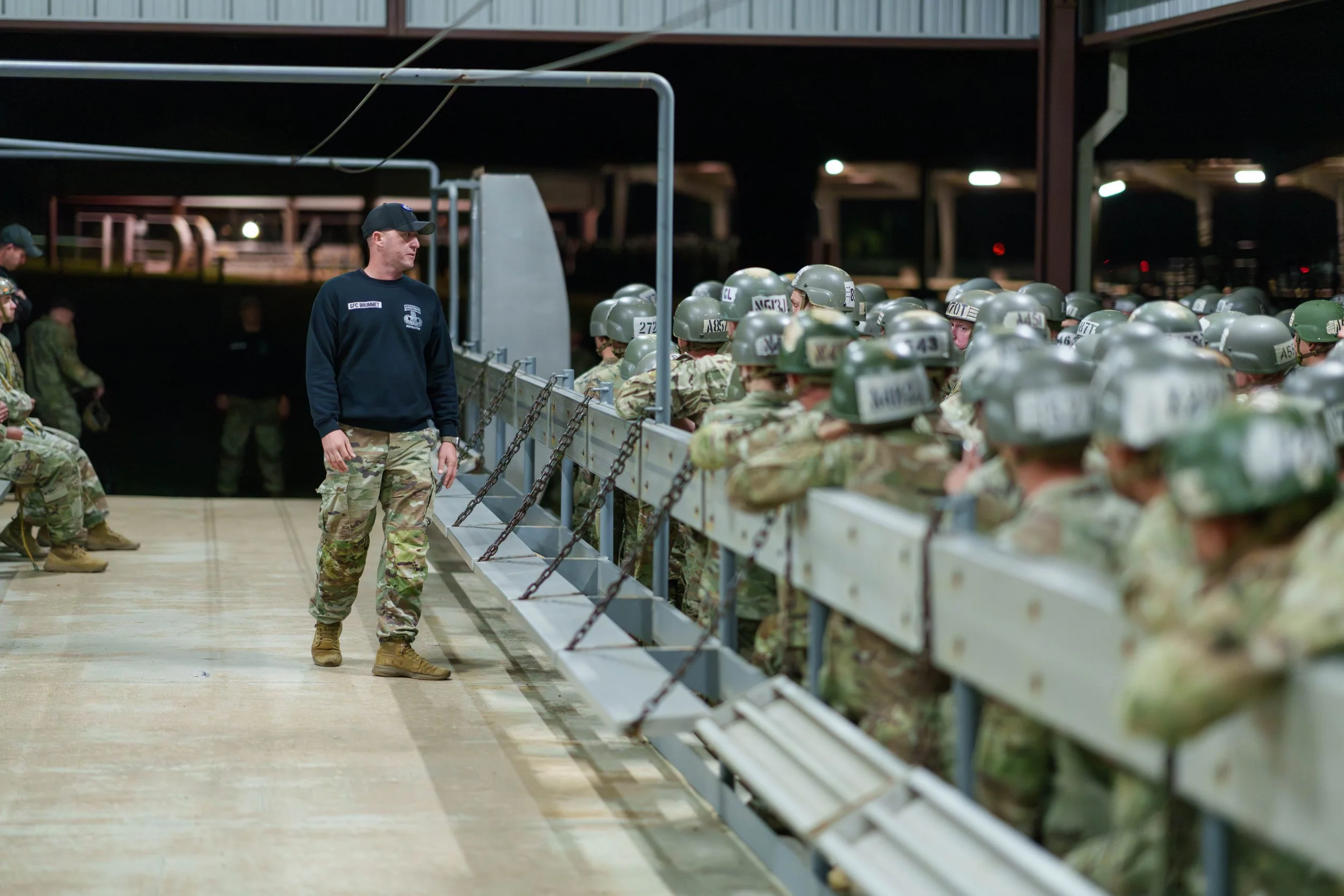 Military personnel in camouflage uniforms and helmets sitting on a bench, with a leader or instructor standing nearby at night.
