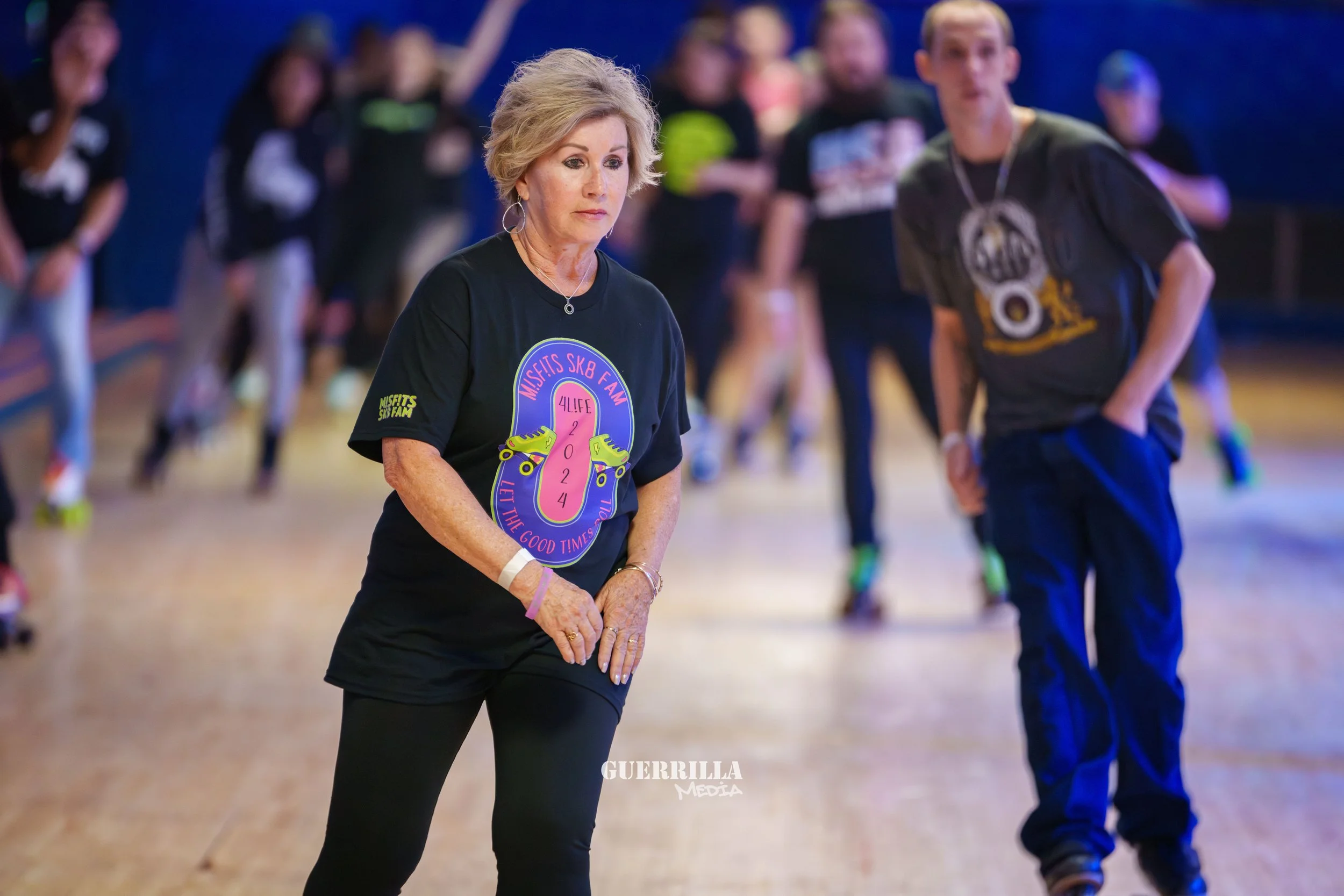 A woman wearing a black T-shirt with colorful roller skate graphics and text, standing in a roller skating rink with other skaters in the background.