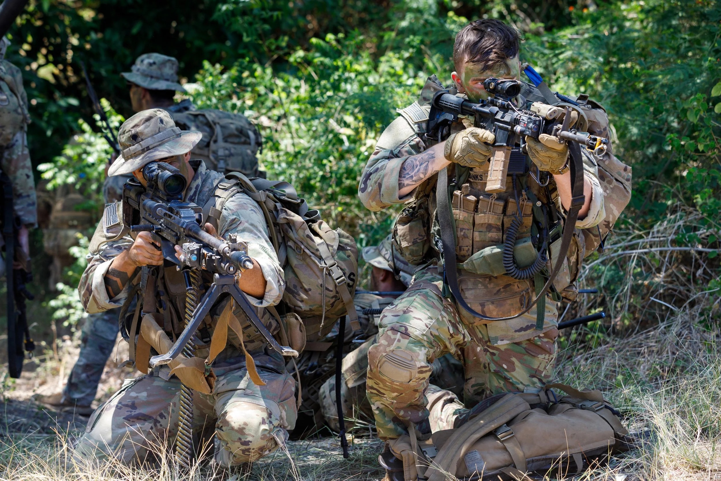 Two soldiers in camouflage uniforms kneeling in a forest, one aiming a rifle, the other looking through a scope on his rifle.