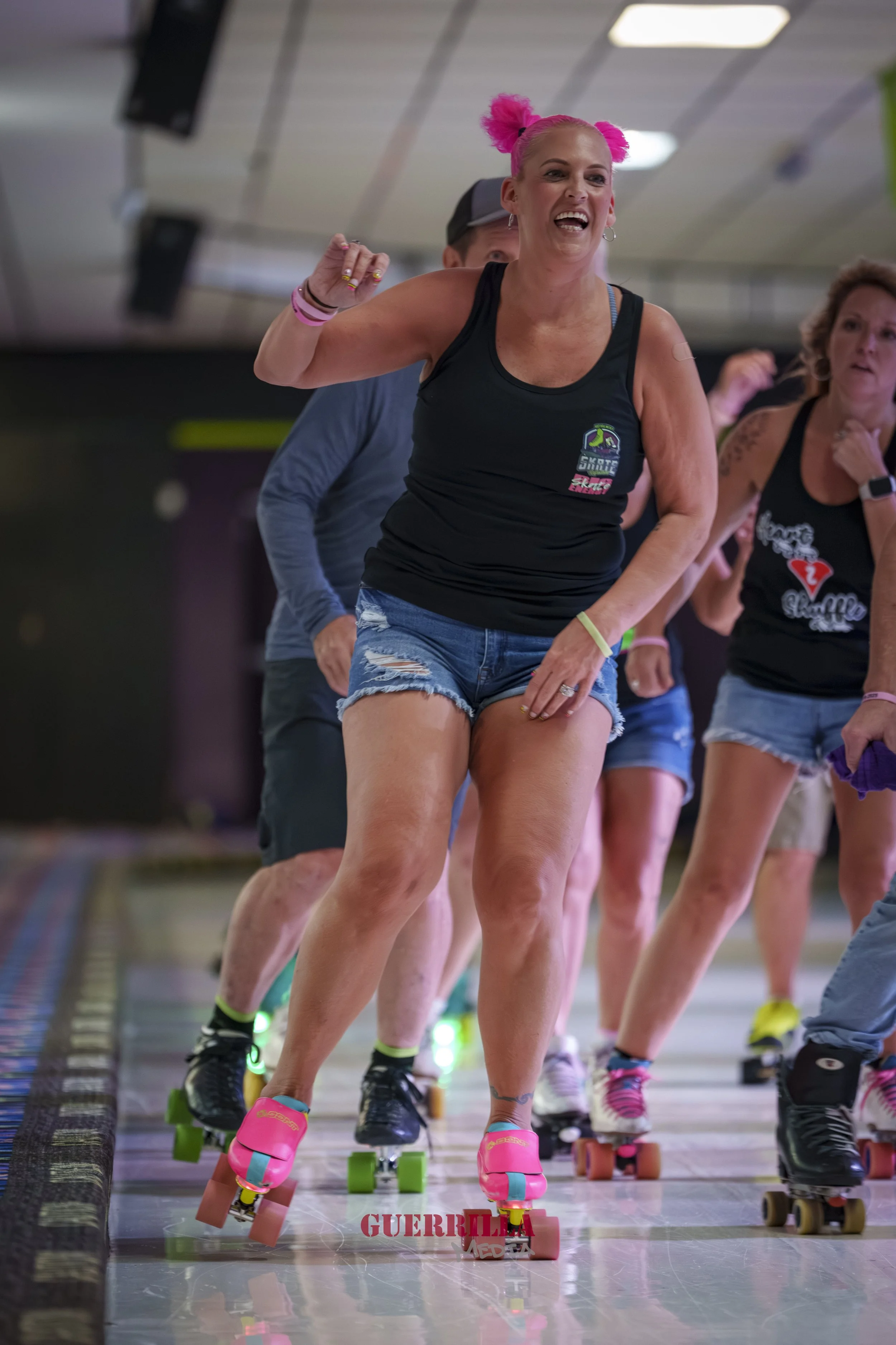 Women roller skating indoors during a skate class or event, with the woman in front wearing a black tank top, denim shorts, and pink skates, raising her fist and smiling.