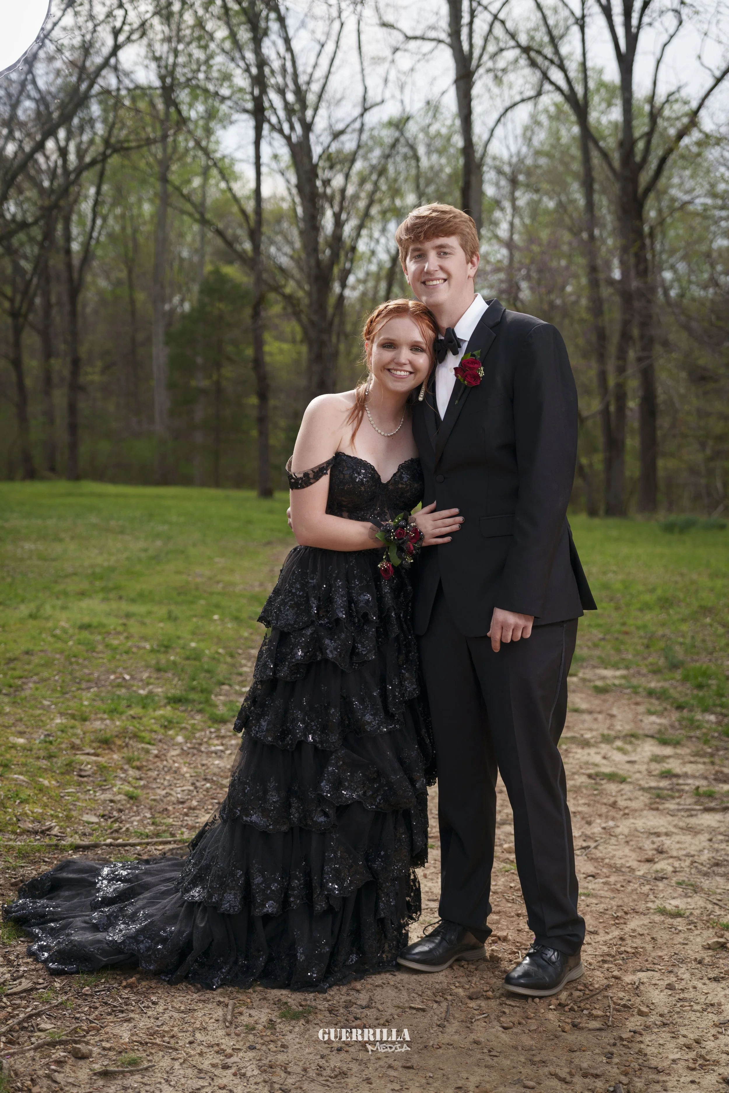 A young couple dressed in formal attire standing together outdoors in a wooded area, smiling at the camera. The woman is wearing a black off-shoulder gown with ruffles and sequins, and the man is in a black tuxedo with a white shirt and black bow tie