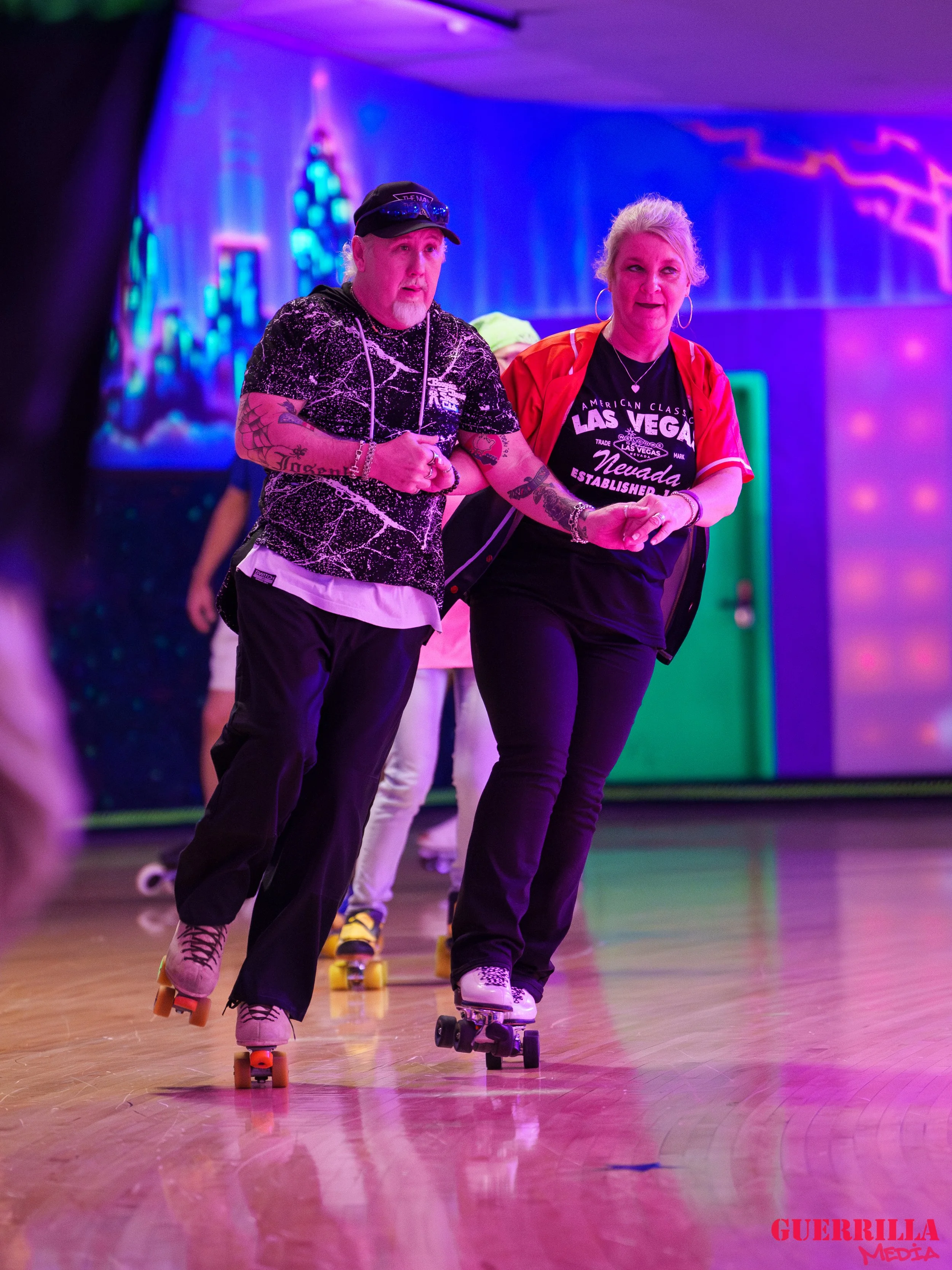 Two women roller skating inside a colorful, neon-lit room, holding hands and balancing on their skates.