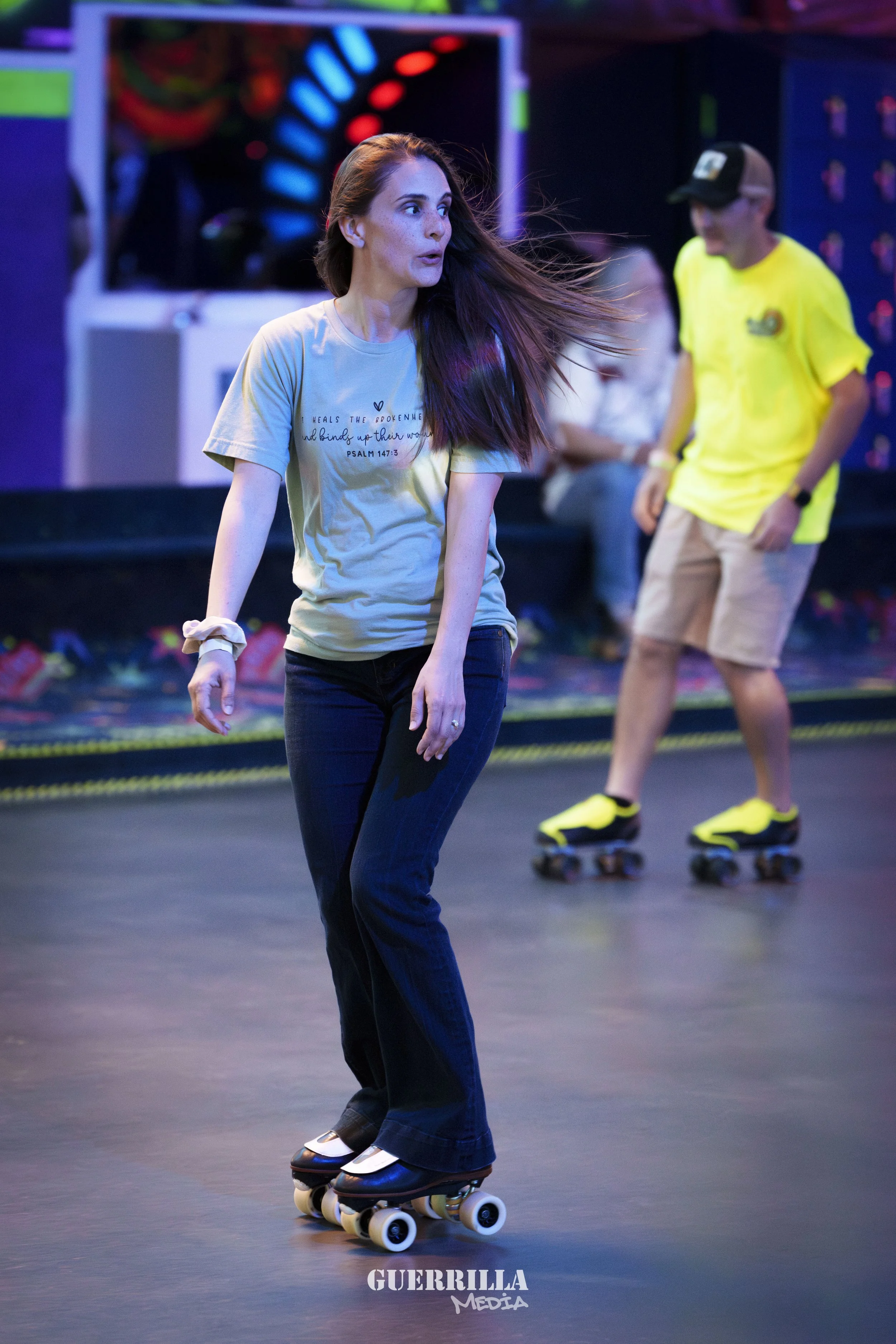 A woman roller skating indoors at an entertainment venue, wearing a light-colored t-shirt and dark jeans, with a man in a bright yellow shirt and shorts skating behind her.