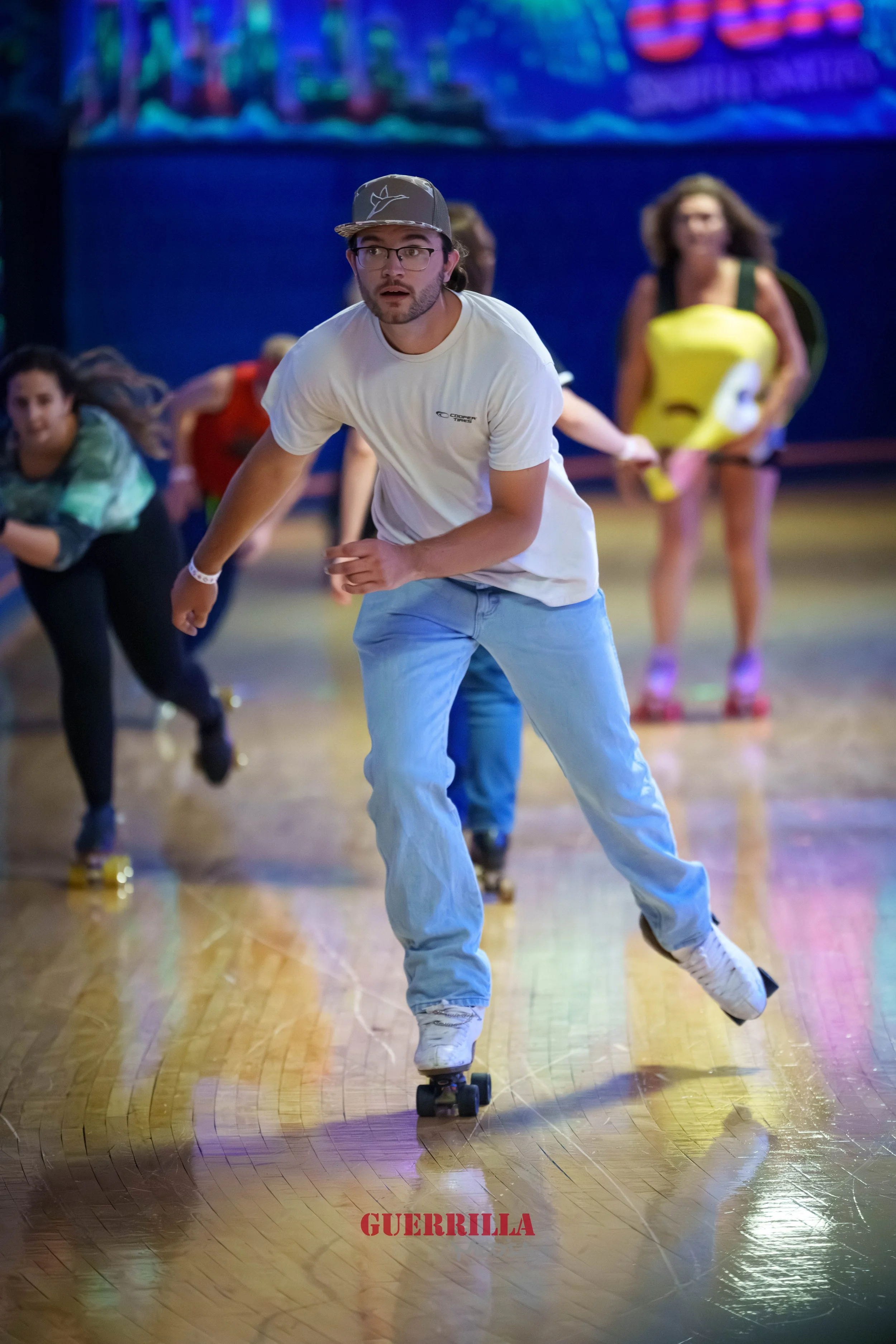 A group of people roller skating indoors, with a man in a white t-shirt and light blue jeans in the foreground. The man is wearing glasses and a gray cap and appears surprised. In the background, a woman is dressed in a yellow costume resembling a ba