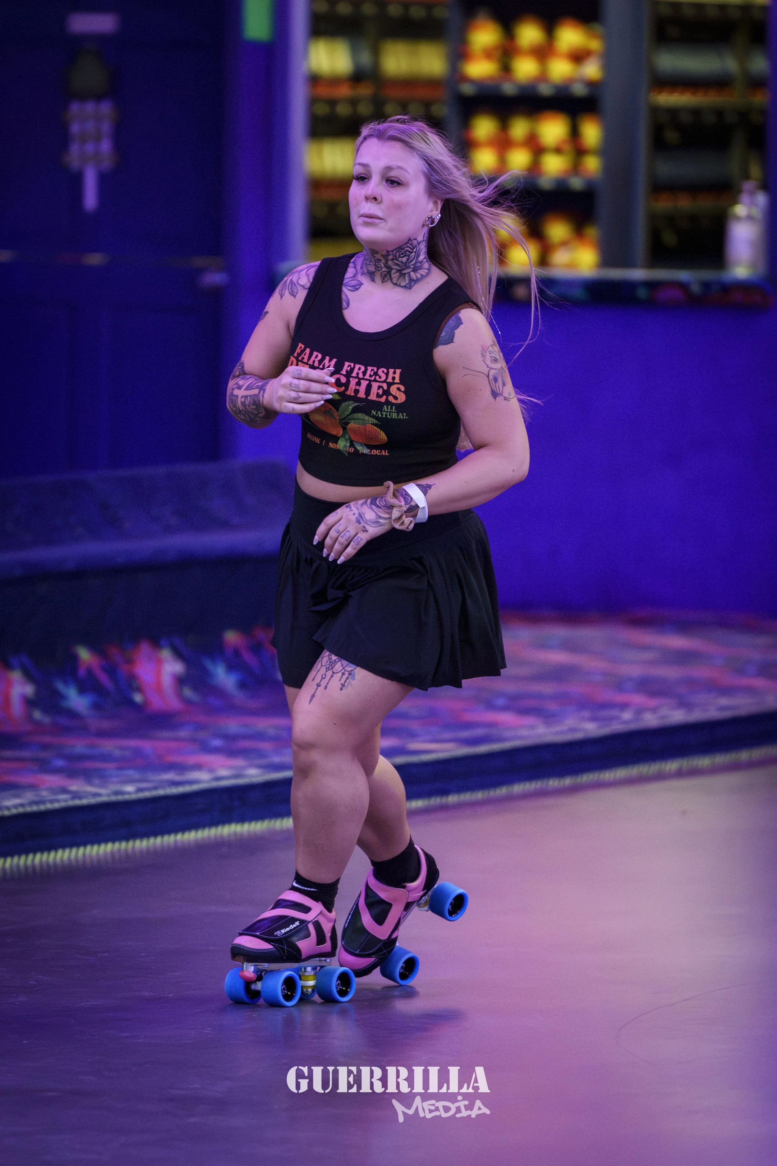A woman roller skating indoors, wearing a black crop top with "Farm Fresh Peaches" text, pink and black roller skates, and sportswear, with tattoos on her arms and neck.