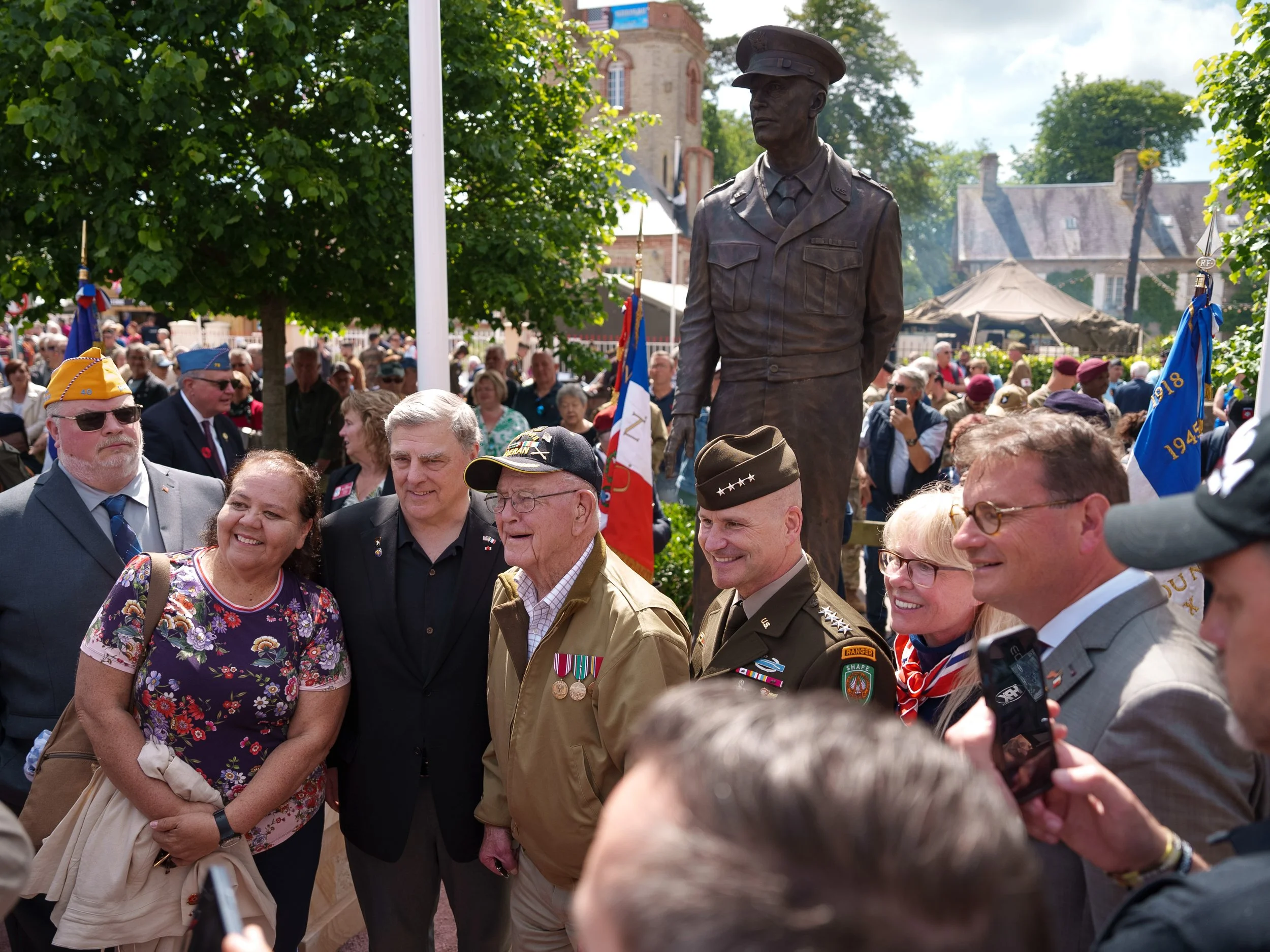 People gathered in front of a military statue during a commemorative event, with flags, trees, and a crowd in the background.