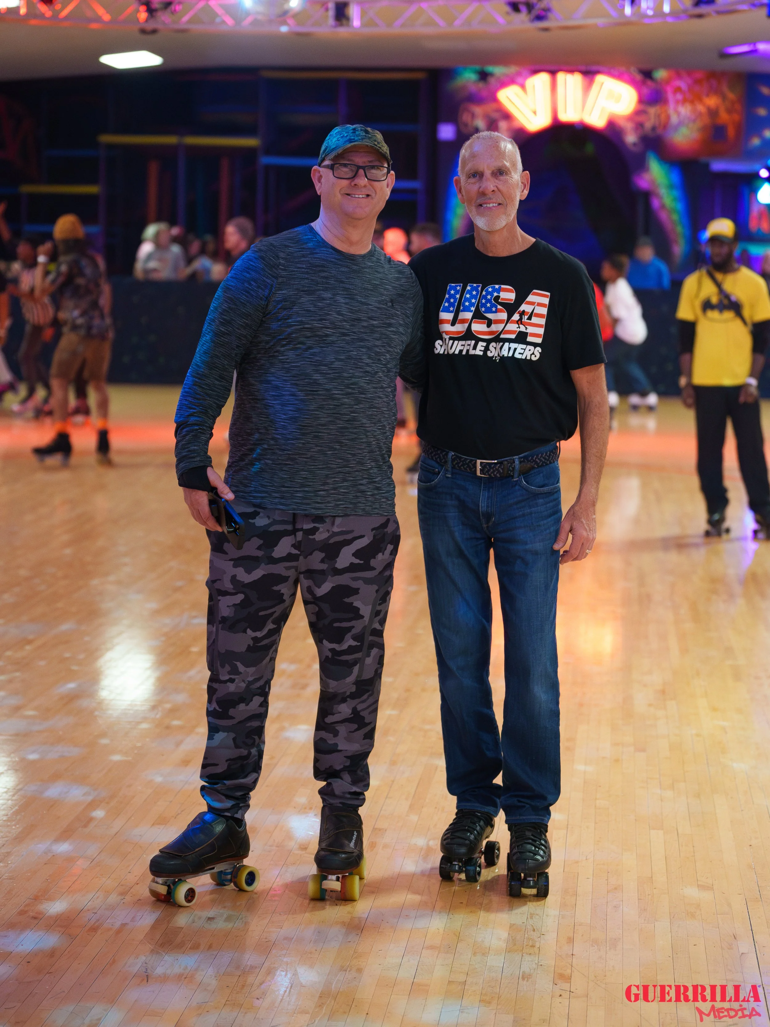 Two men with roller skates at an indoor roller rink, smiling for the camera. The background shows colorful lighting, other skaters, and a neon sign that says 'VIP'.