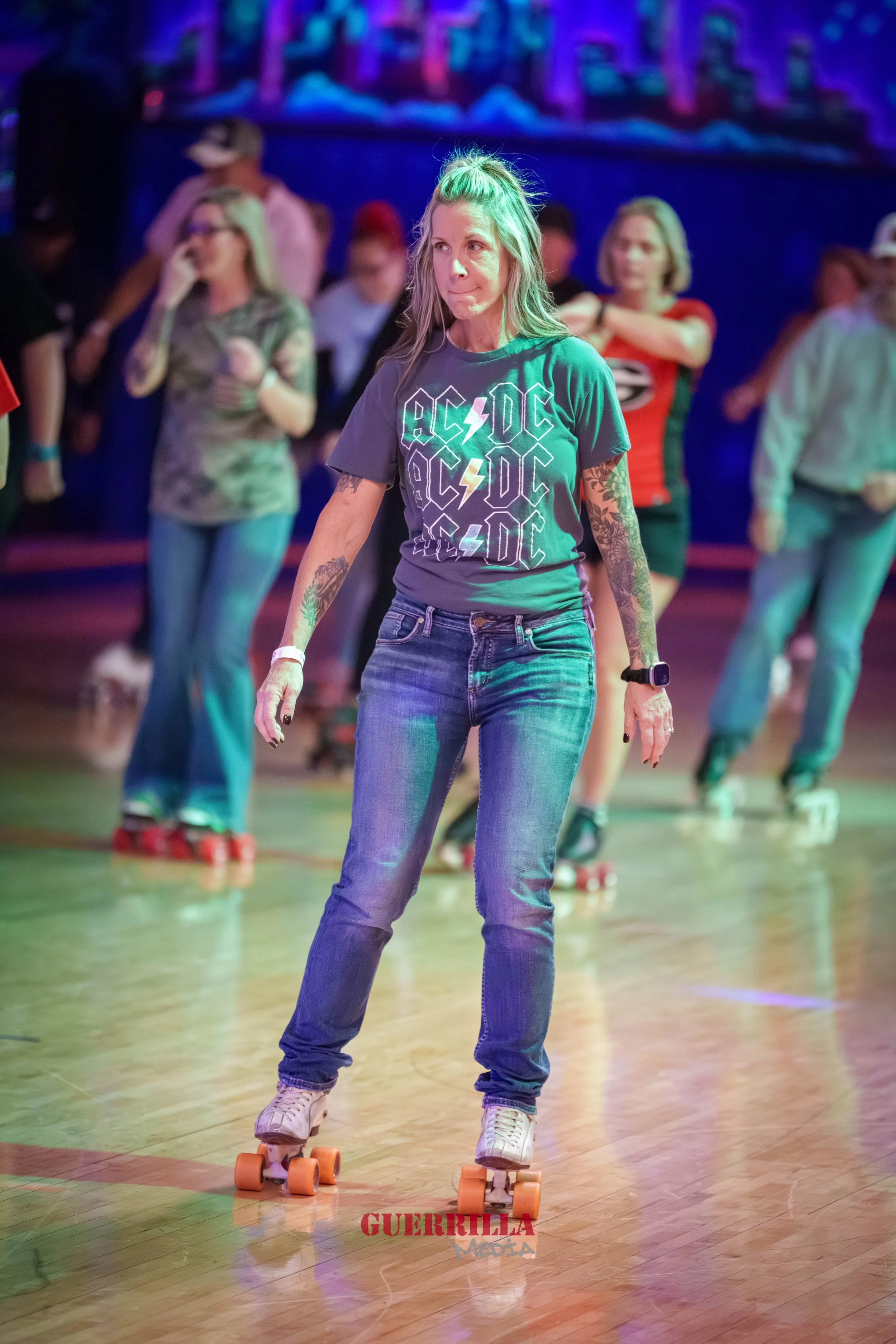 A woman roller skating indoors at a roller rink, wearing a gray AC/DC t-shirt and jeans, with tattoos on her arms, and surrounded by other roller skaters in the background.