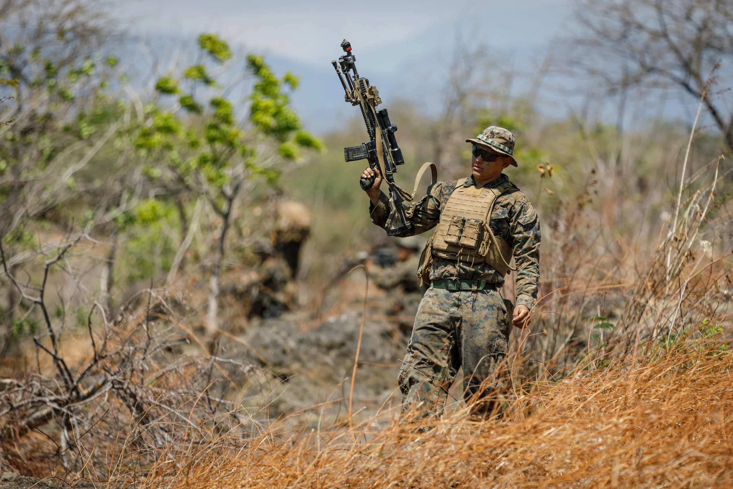 A soldier in camouflage uniform, wearing a tactical vest and hat, standing amidst dry grass and trees, holding a rifle in a firing stance outdoors.