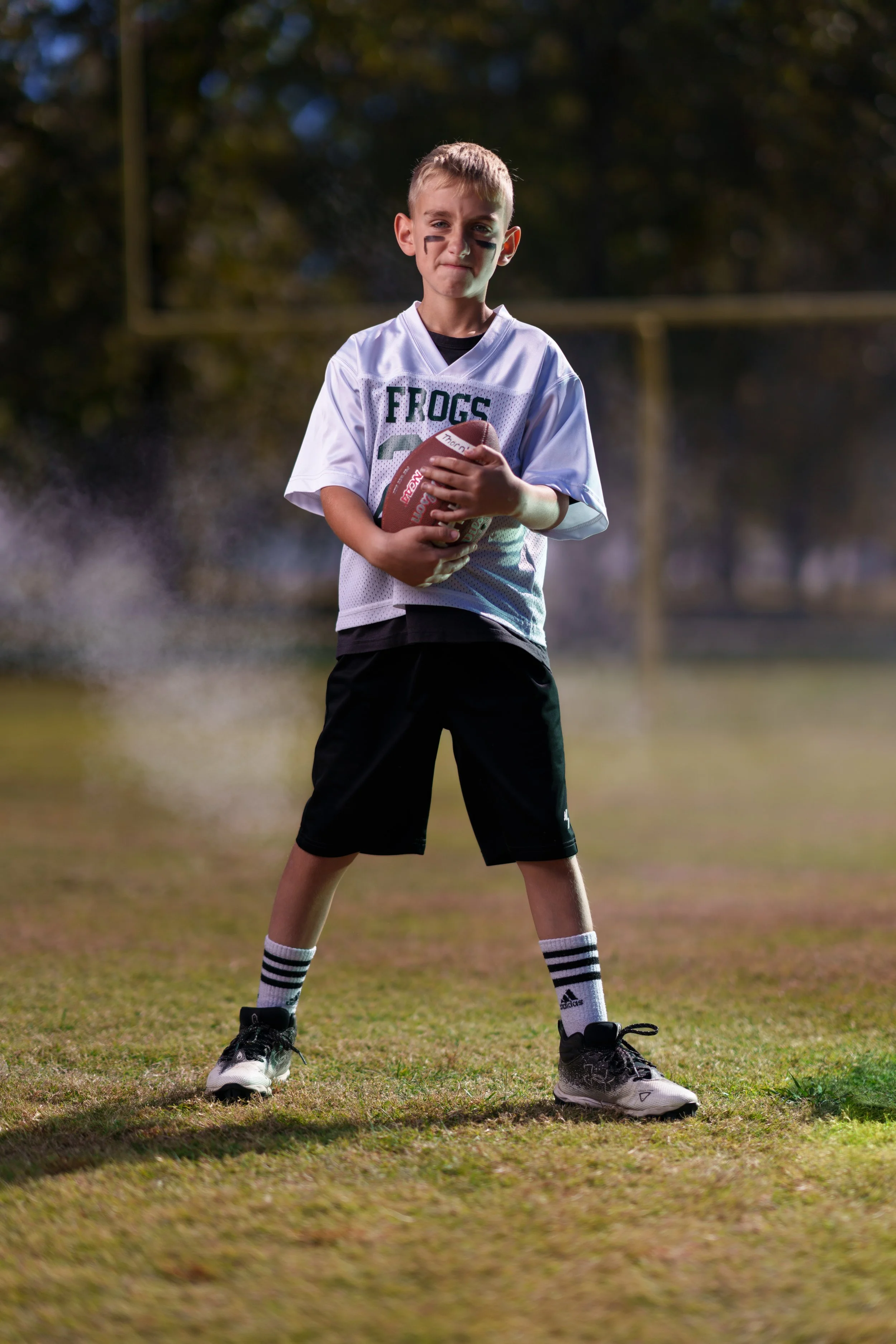 Young boy on football field holding a football, wearing a football jersey, shorts, athletic shoes, and striped socks, with face paint and a serious expression.