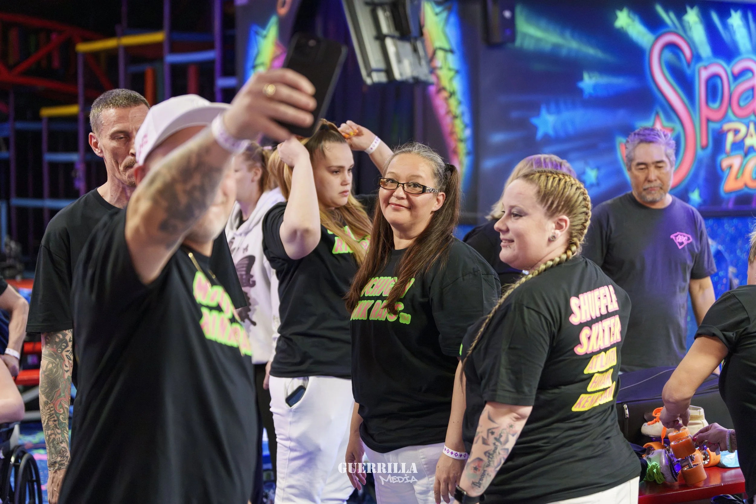Group of people at an indoor roller skating event, some taking selfies, with roller skates on a table and a colorful, vibrant background with the words 'Space' and 'Zoo' visible.