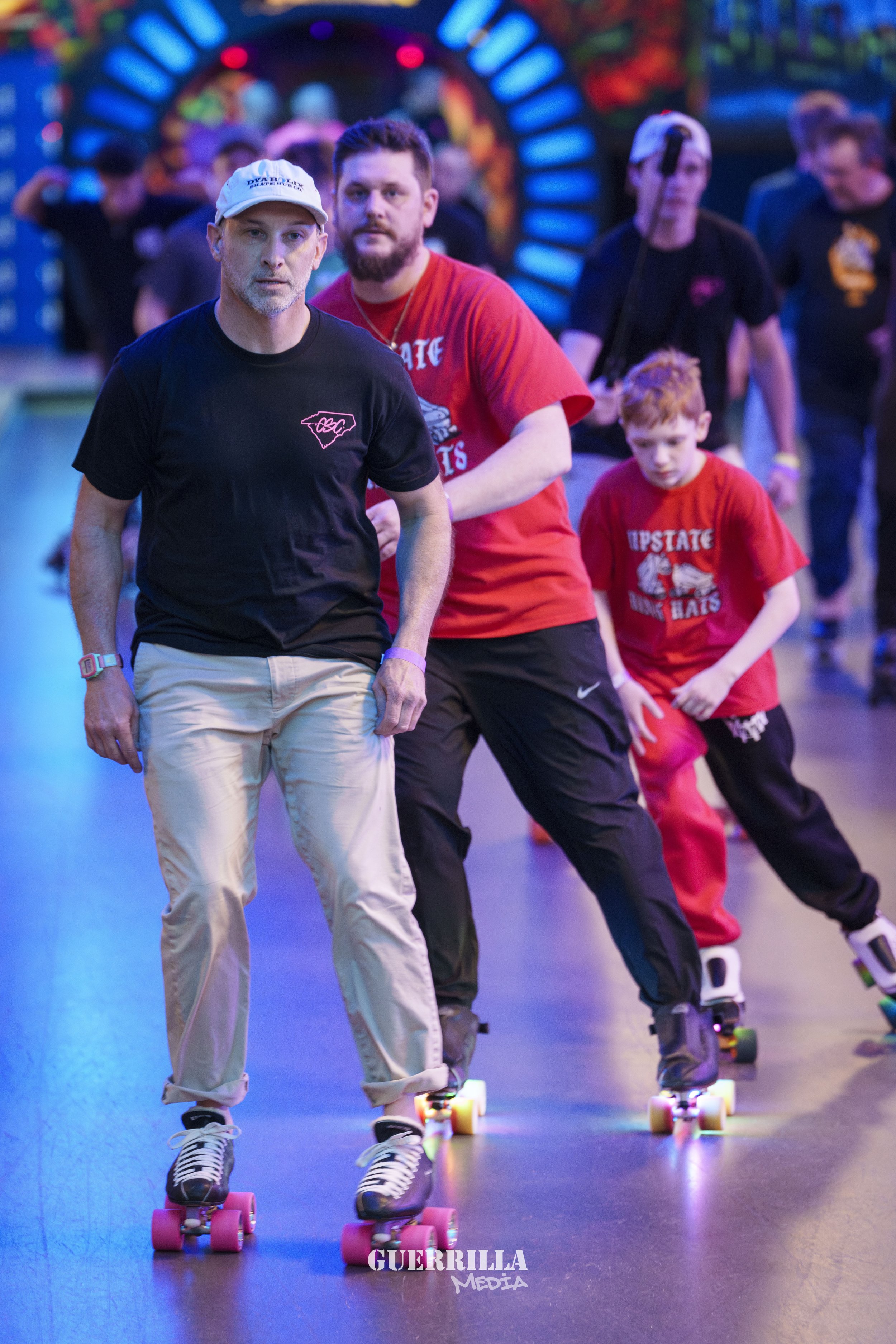 People roller skating indoors, with a colorful, neon-lit background and a man in a black shirt and beige pants leading a child and another man, all wearing casual athletic clothing, with the child wearing a red shirt.
