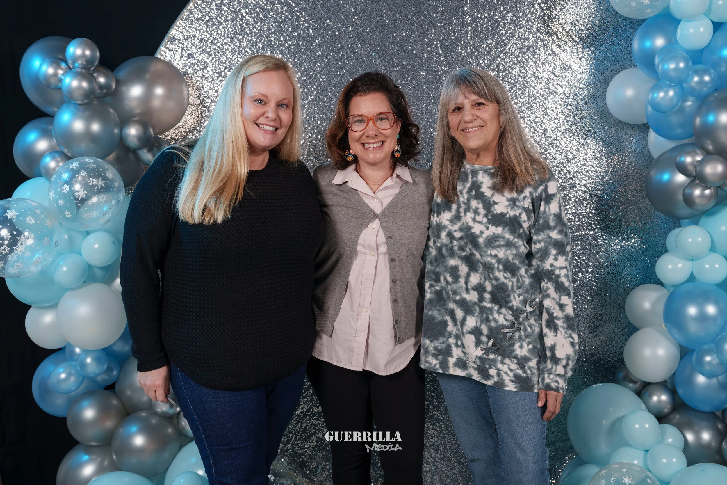 Three women standing together in front of a silver and blue balloon arch and glittery backdrop, smiling for the camera.