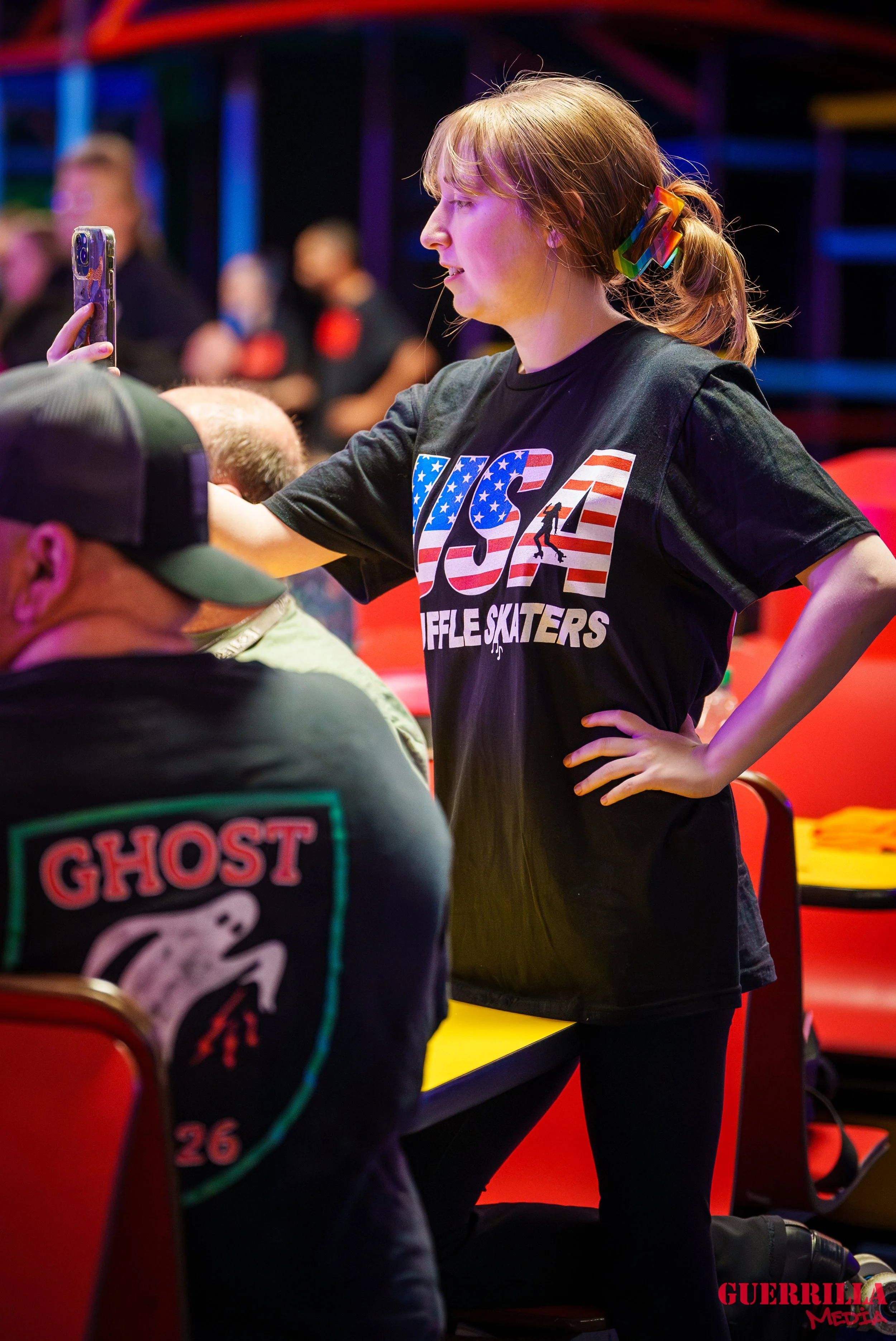 A young woman with red hair tied back with a rainbow-colored hair clip, wearing a black T-shirt with an American flag pattern and the words 'USA' and 'Rifle Skaters,' is standing with one hand on her hip and taking a selfie at an indoor skatepark or 