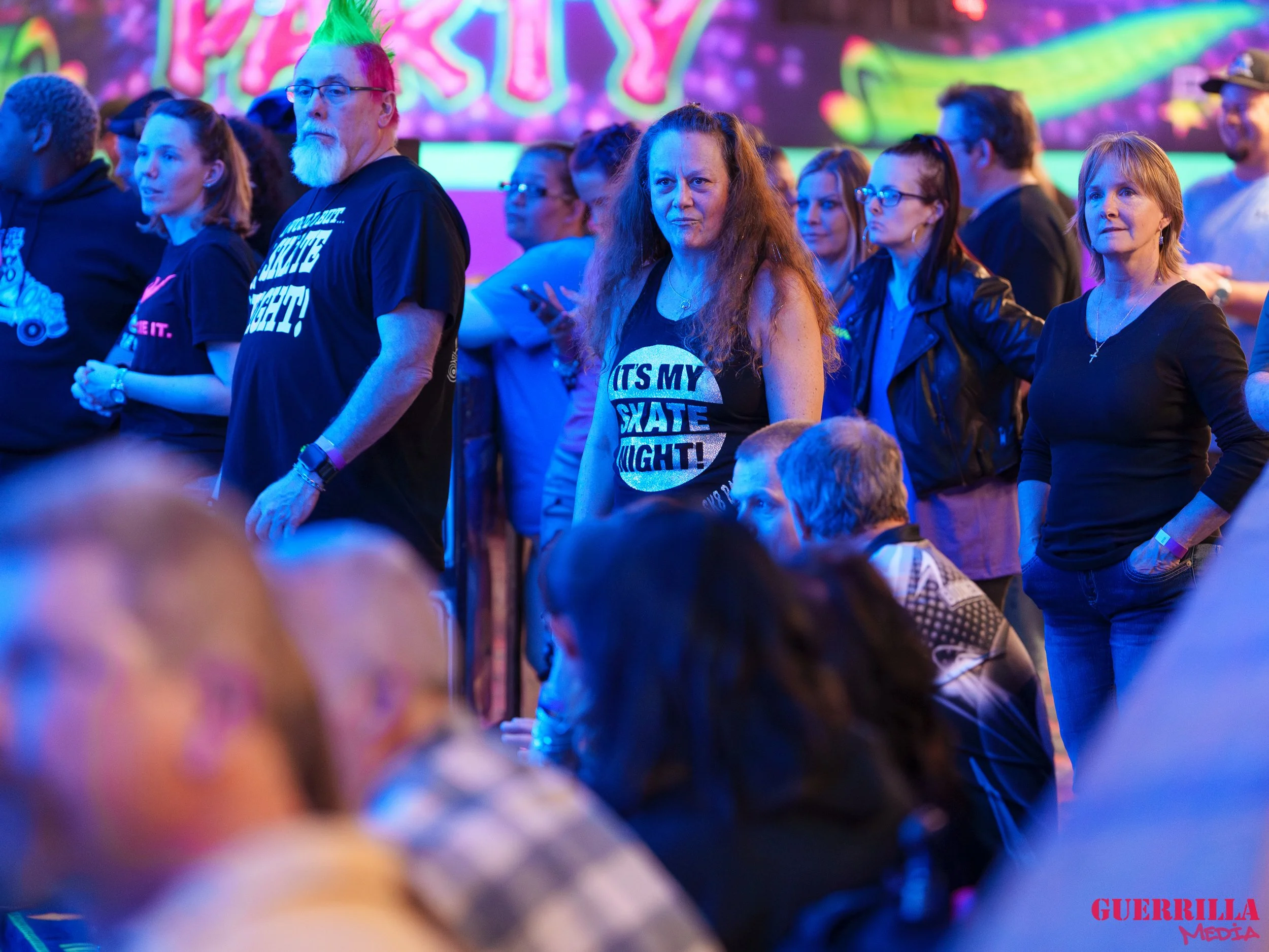 People standing and sitting at an indoor event with neon-colored lighting and a brightly lit backdrop.
