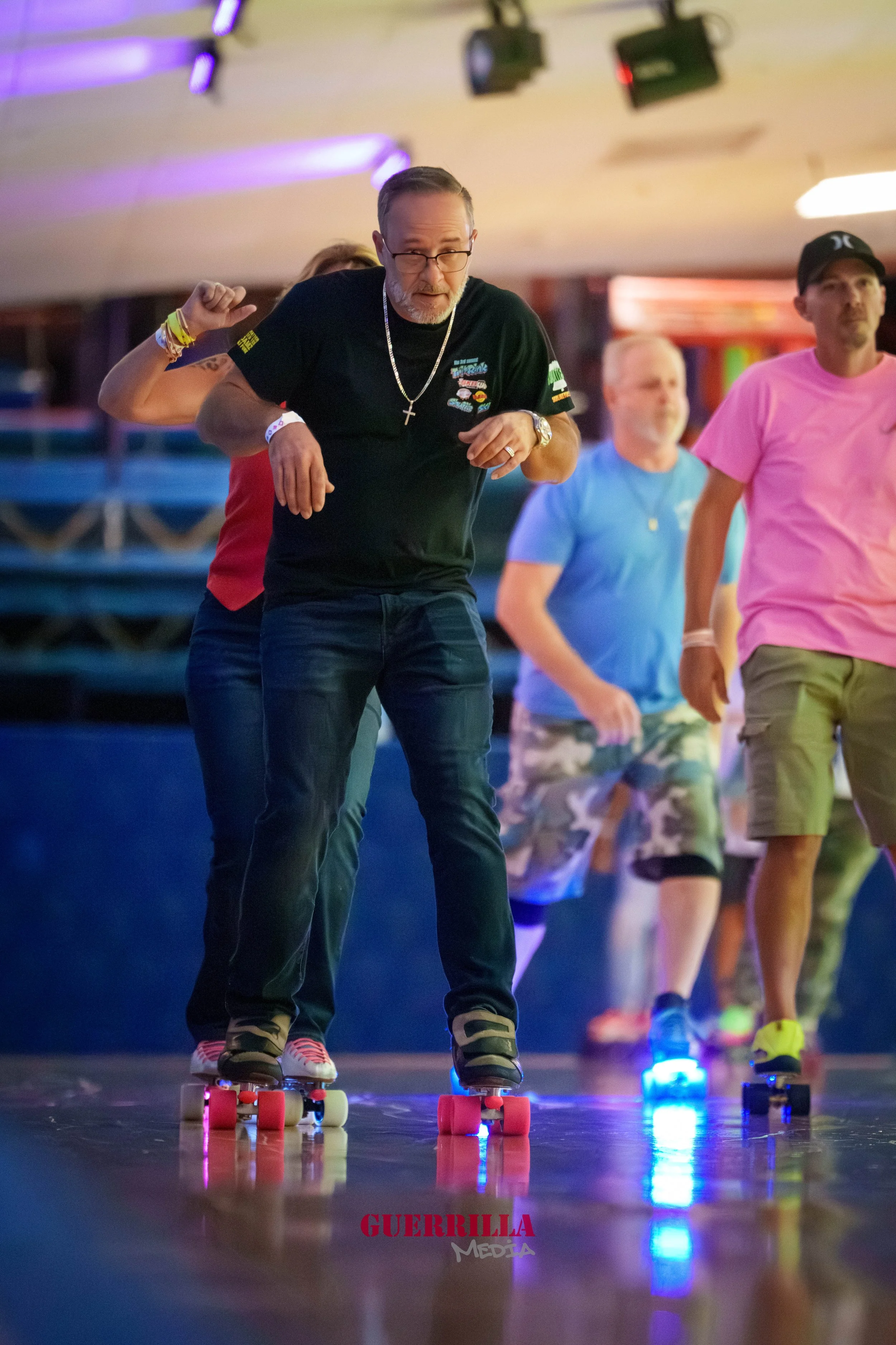 People roller skating indoors, with one man in black shirt and glasses leading the group, colorful lighting, and a reflective wooden floor.