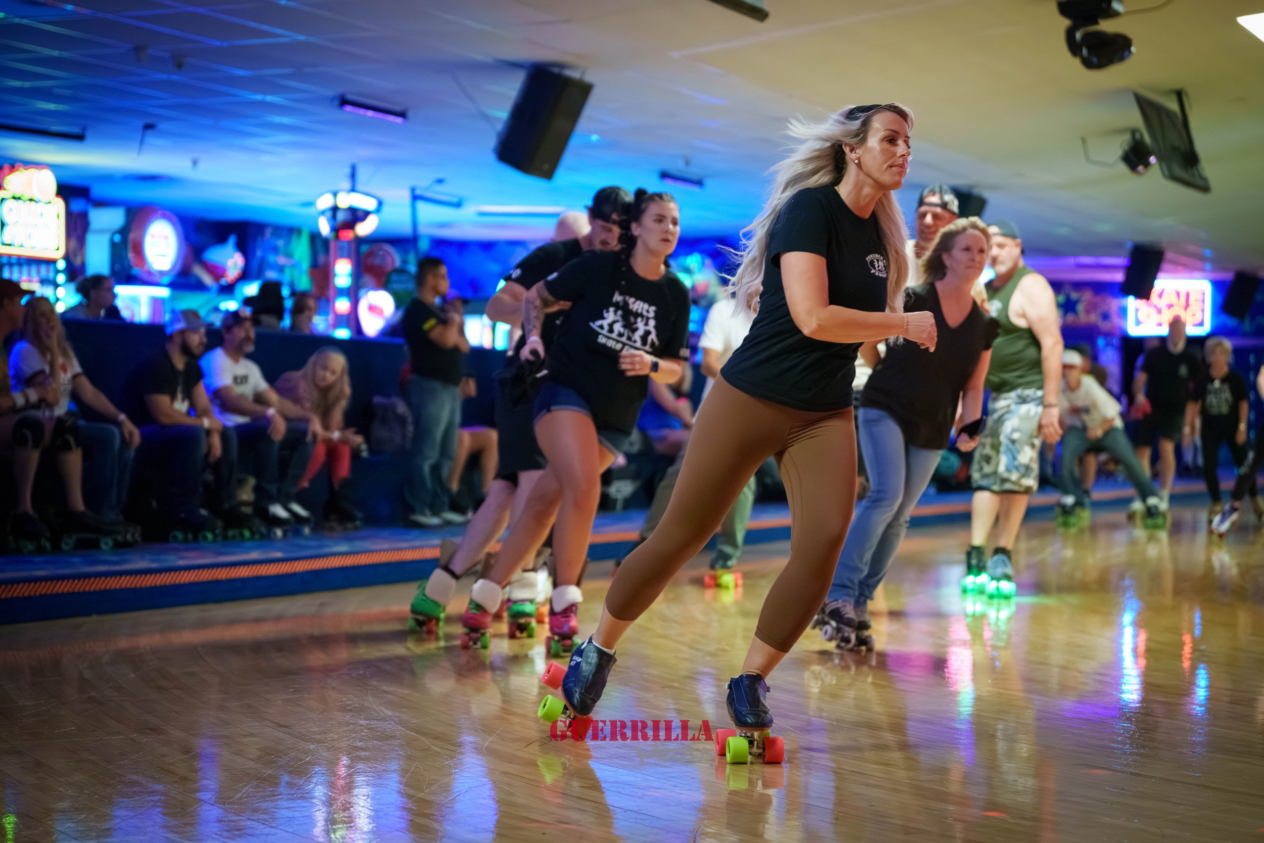 Group of people roller skating indoors under colorful neon lights, with some seated spectators watching.