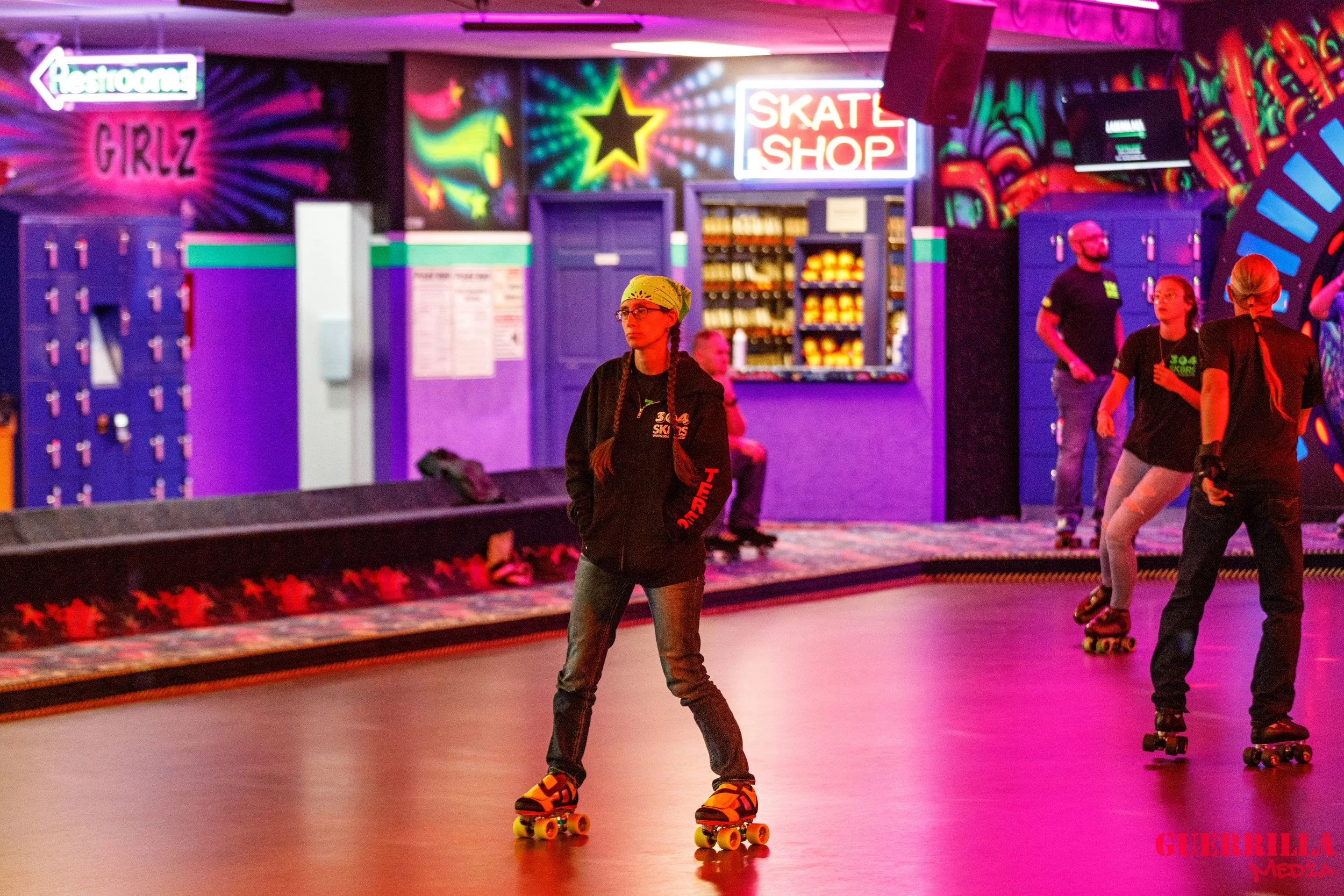 People roller skating inside an roller-skating rink illuminated by vibrant, colorful neon lights. The background features lockers, signs for a snack shop and restrooms, and vivid wall art.